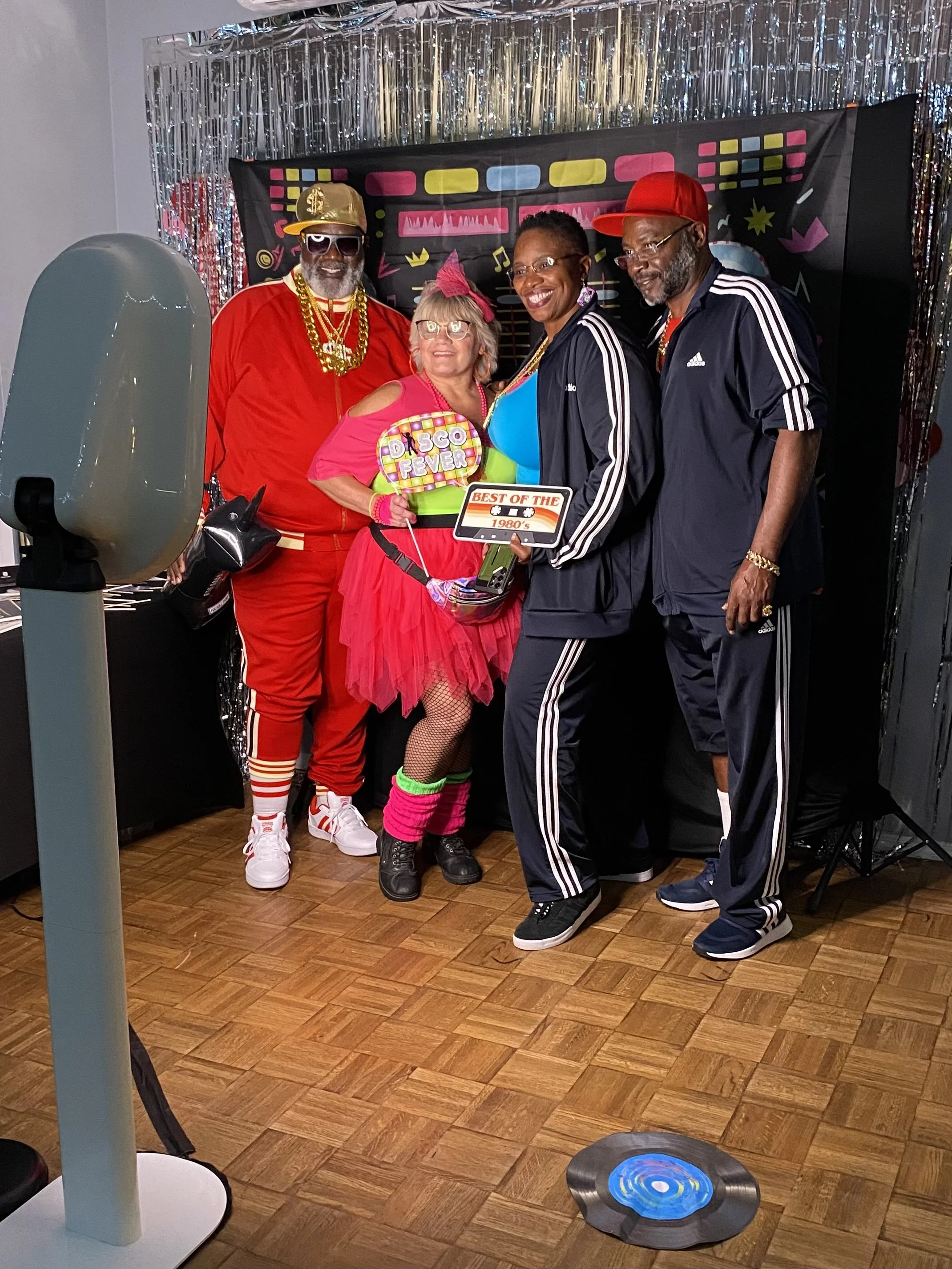 Four adults dressed in colorful and sporty outfits at a 1980s-themed party, standing together and smiling for a photo, with a shiny silver fringe backdrop and a decorative sign that reads "Best of the 1980s."