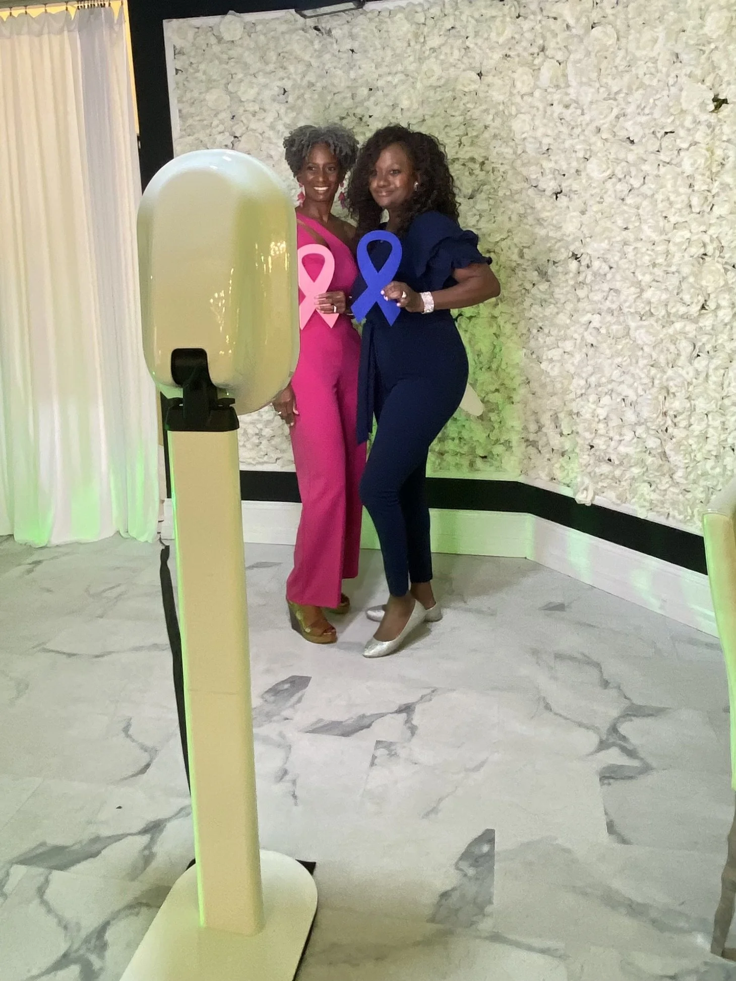 Two women standing close together, holding pink and blue awareness ribbons, in a room with white floral wall backdrop and white marble flooring. One woman wears a bright pink outfit, the other a dark blue outfit, both smiling and facing the camera.