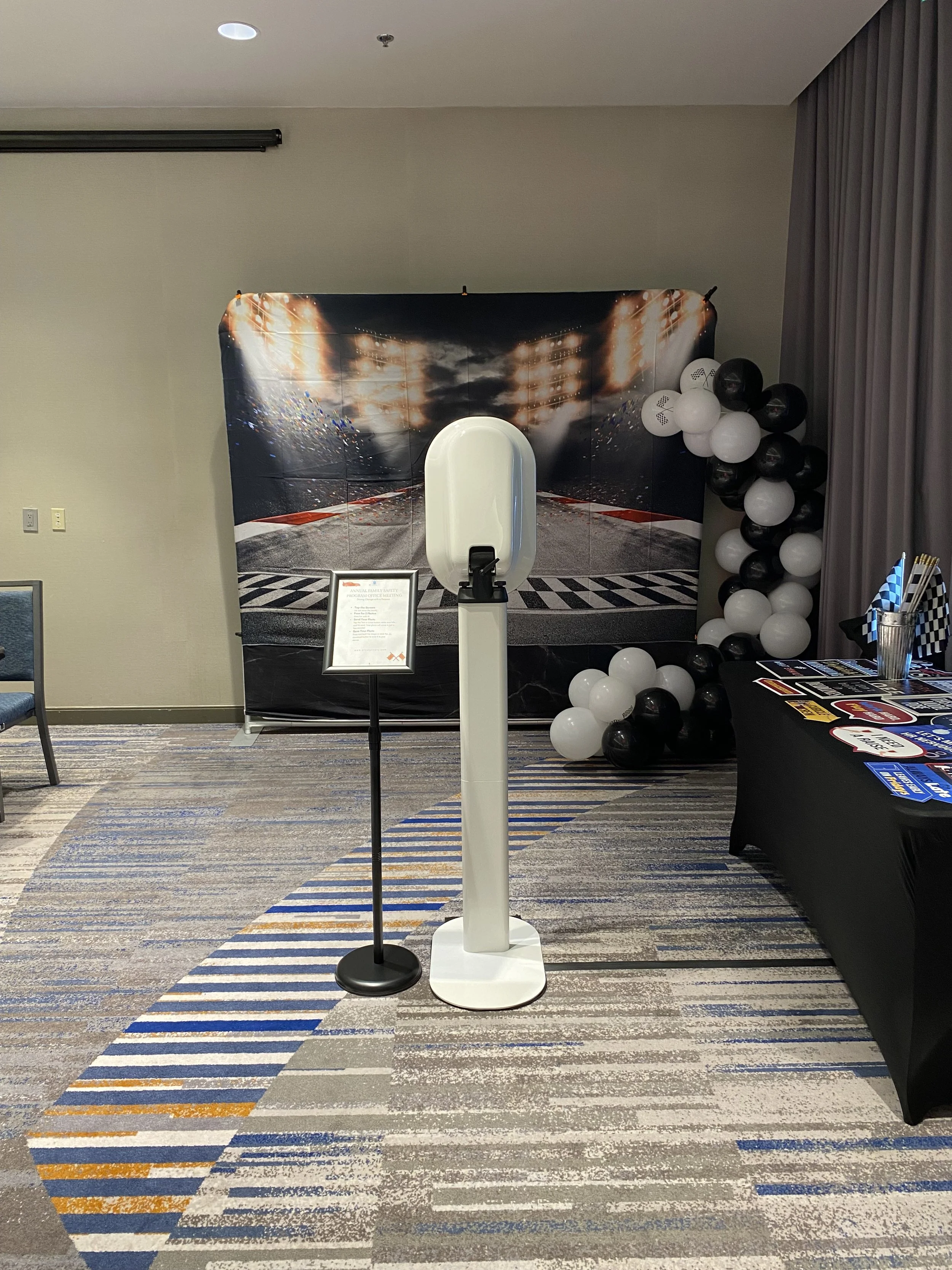 Photo of a photo booth setup with a race track backdrop, a black and white risk-themed balloon arrangement, and a table with race-themed decorations in an indoor space.