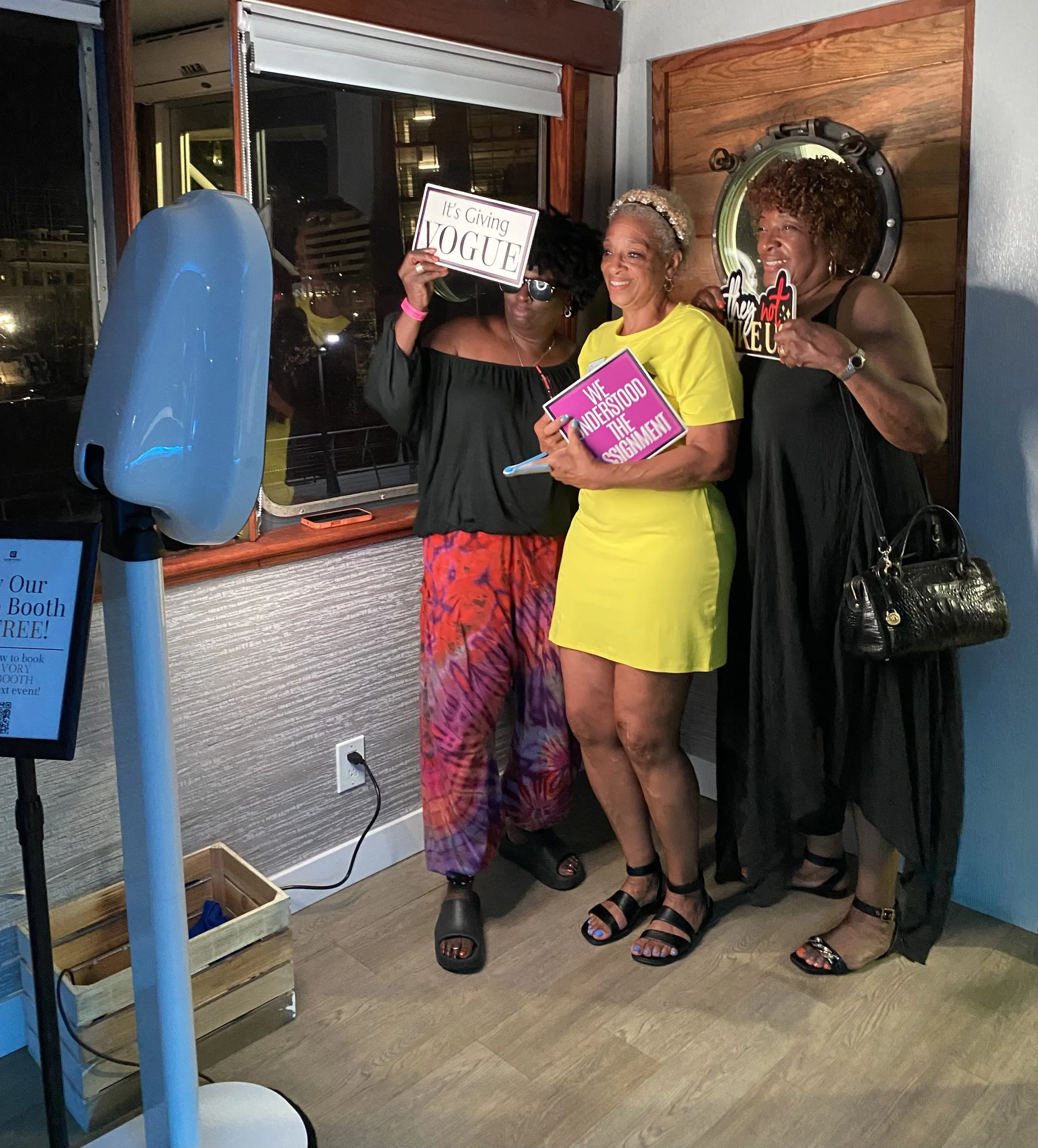Three women standing inside a room near a window, holding signs related to a book event. The woman on the left, wearing a black off-the-shoulder top and sunglasses, is holding a sign that reads 'It's Giving YOGUE'. The woman in the middle, wearing a 