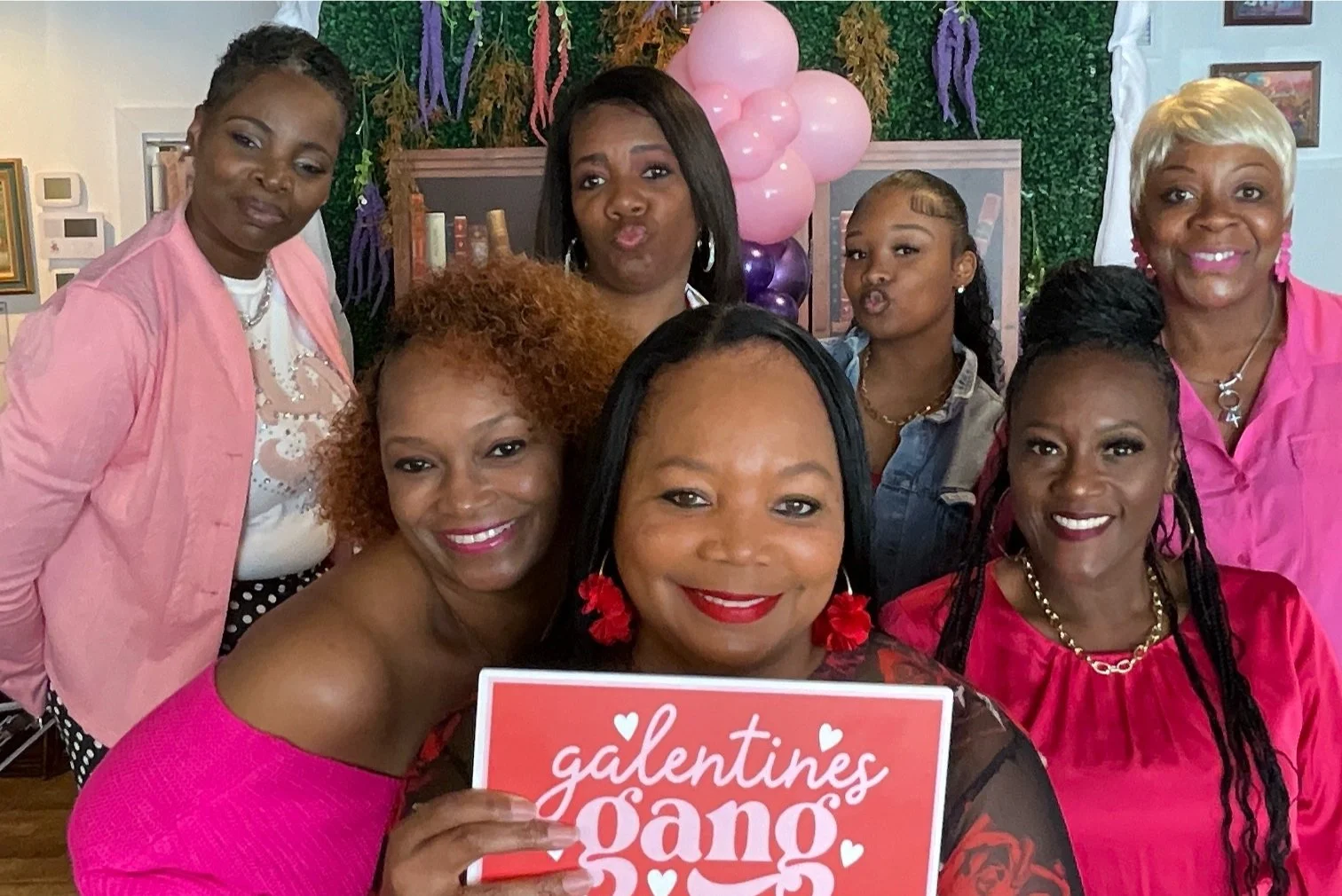 A group of women celebrating Valentine's Day, holding a sign that says 'galentine's gang', with balloons and decorations in the background.