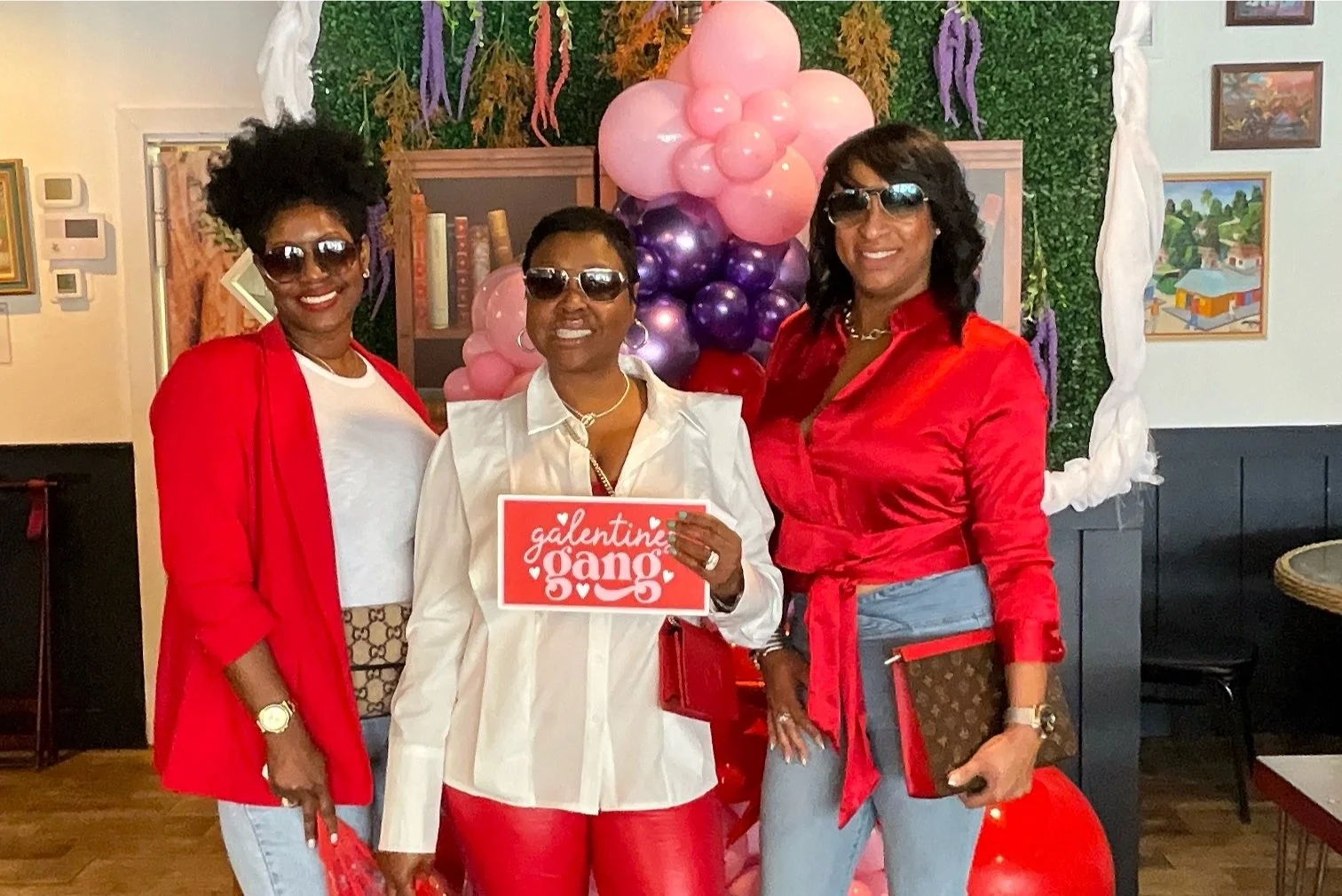 Three women with sunglasses posing together at a Valentine's Day celebration, with pink, purple, and red balloons arranged in a heart shape in the background, in an indoor setting.