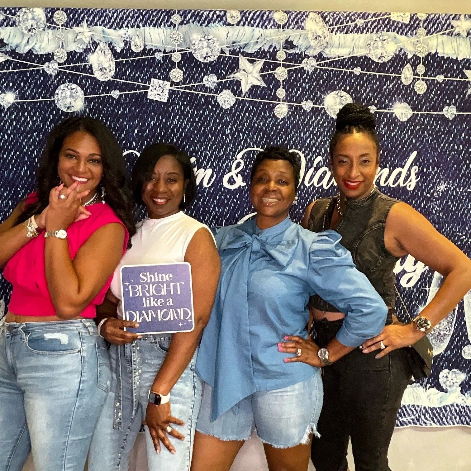 Four women stand together in front of a decorated backdrop, smiling. They are dressed in casual and semi-formal outfits, some wearing jeans and others in a blouse or sleeveless dresses. One woman is holding a sign that says 'Shine Bright like a DIAMO