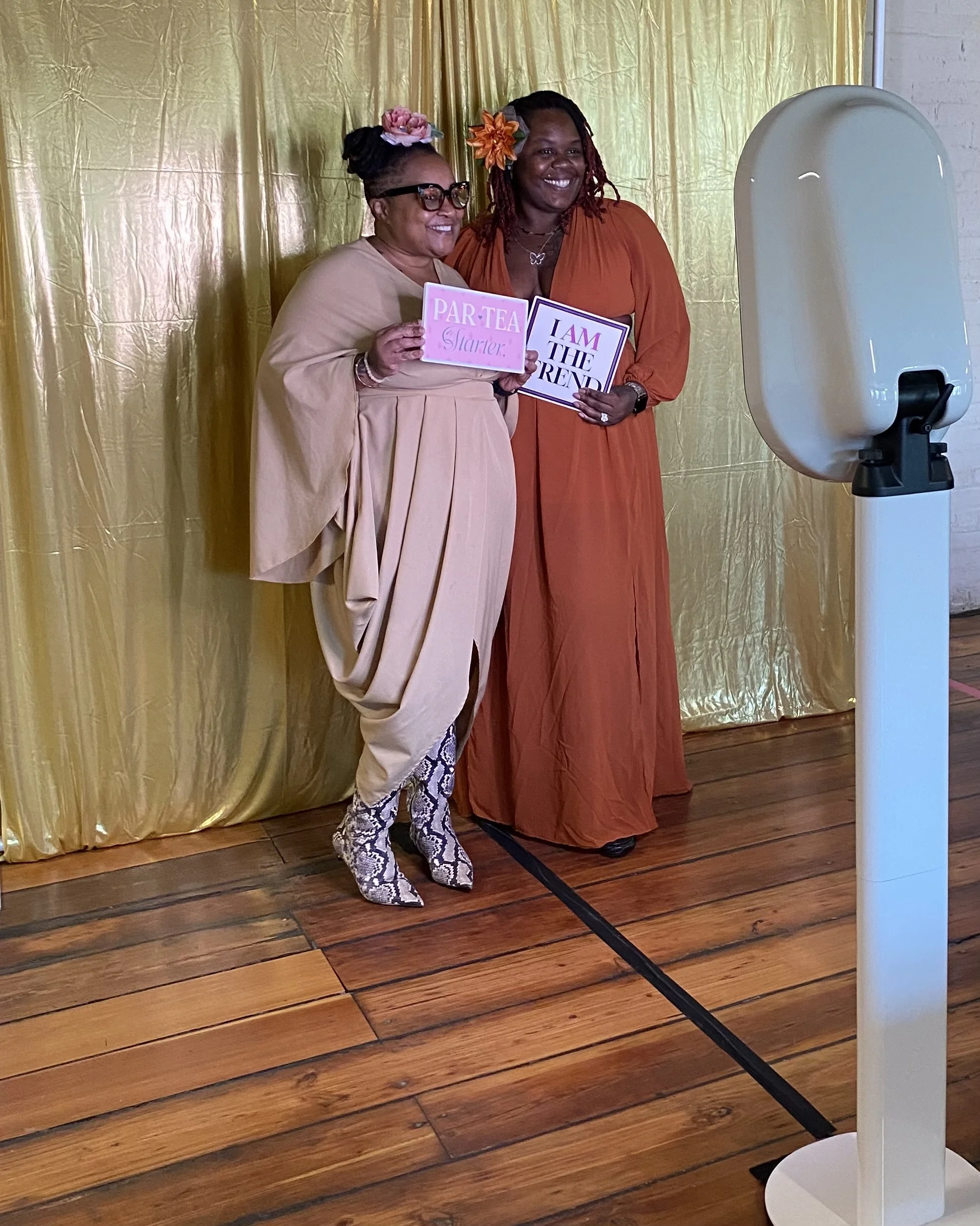 Two women smiling and holding signs, standing in front of a gold curtain. One woman wears a beige outfit with floral headwear and snake print pants. The other wears an orange dress with a flower in her hair. There is a large white photobooth or skin scanner to the right.