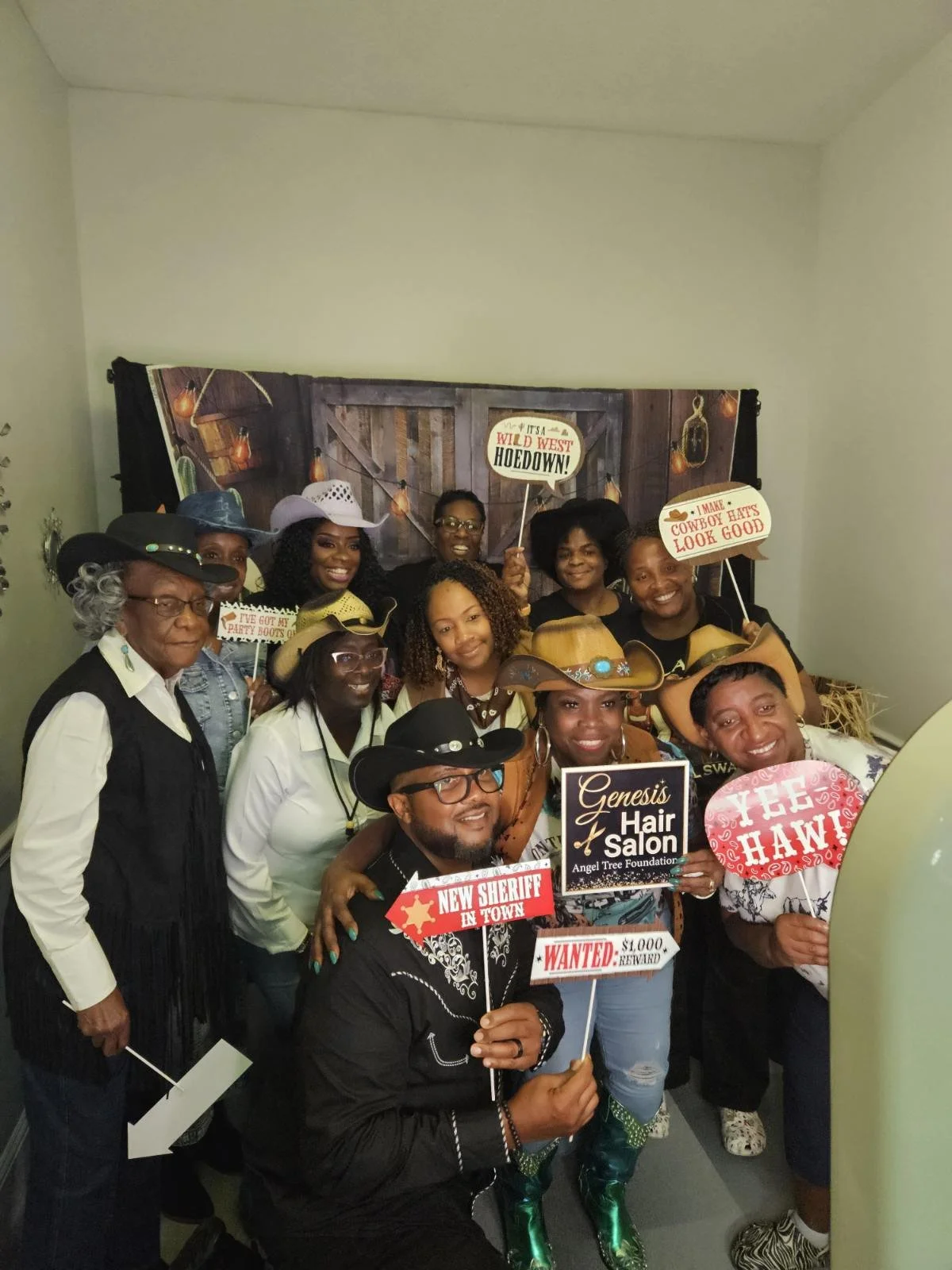 Group of people dressed in cowboy and western themed clothing at a photo booth, holding themed signs, with a rustic western background.