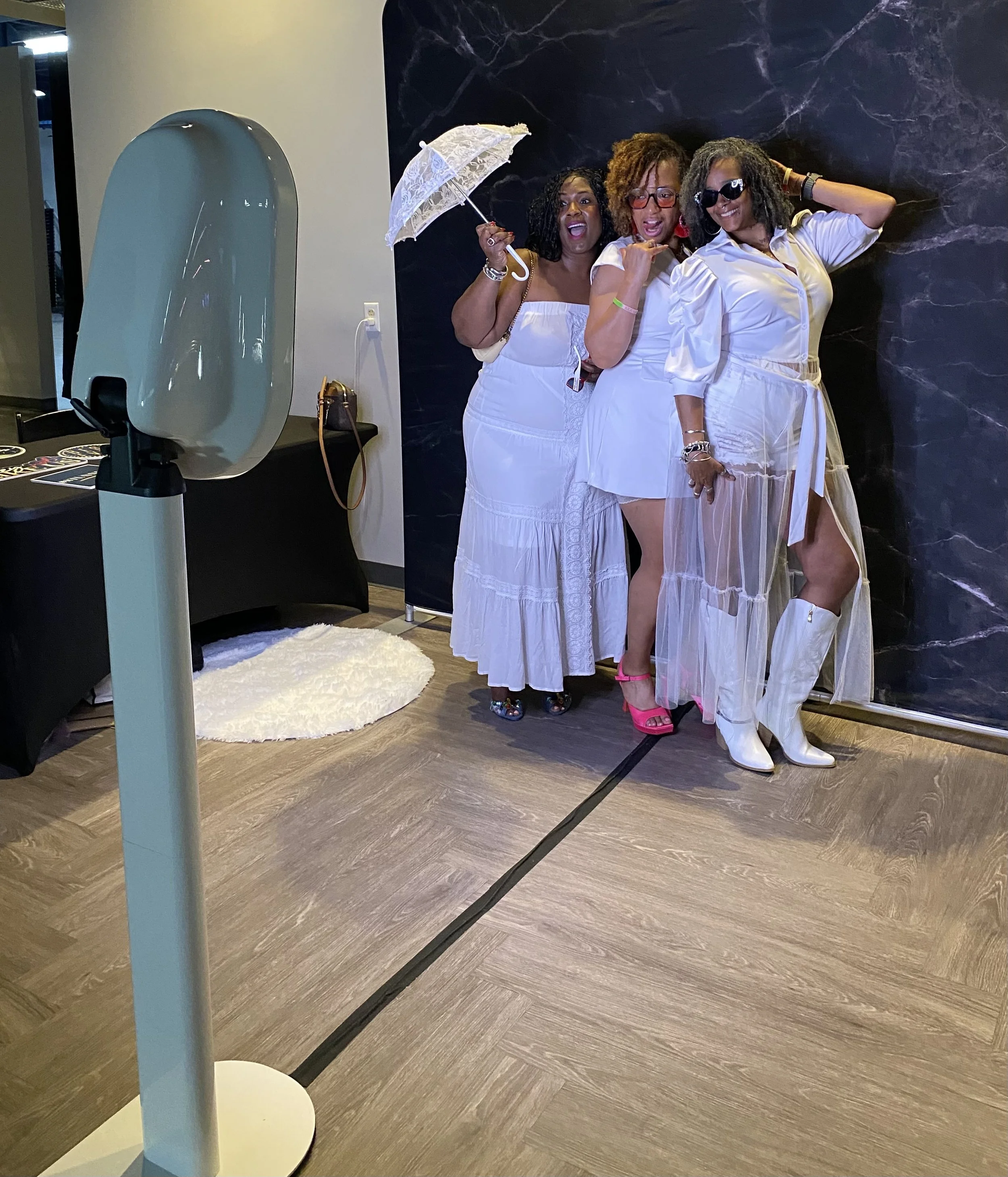 Three women posing in front of a black marble wall, with a white floor, a white lace umbrella, using PIXSBYIVORY PHOTO BOOTH.