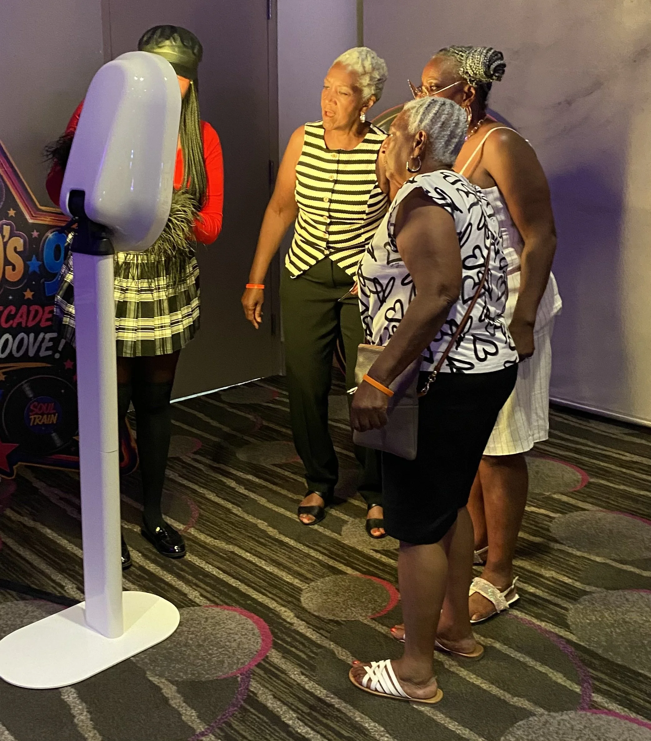 A group of women, dressed in casual and stylish clothes, stands in a semi-circle around a white tracking device with a stand in an indoor setting with patterned carpet. They appear to be in conversation or waiting for something.