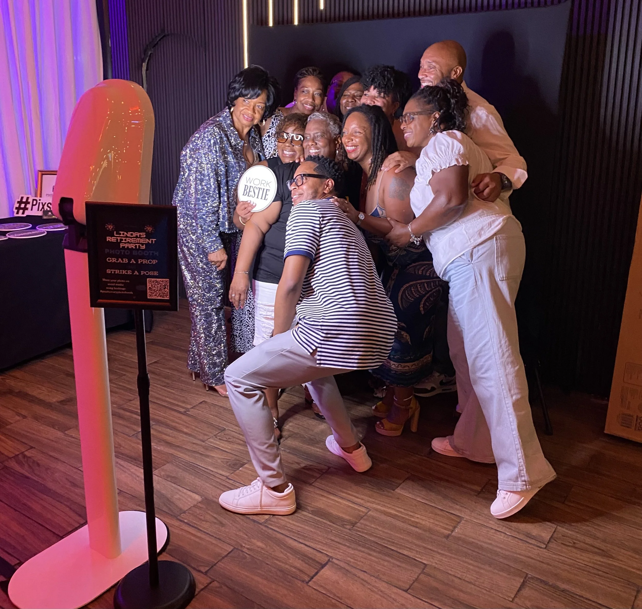 Group of people hugging and smiling in a photo booth at a retirement party. There are signs and props nearby, including one that says 'WORK BESTIE' and another advertising a photo booth. The setting appears to be a decorated indoor event space with w