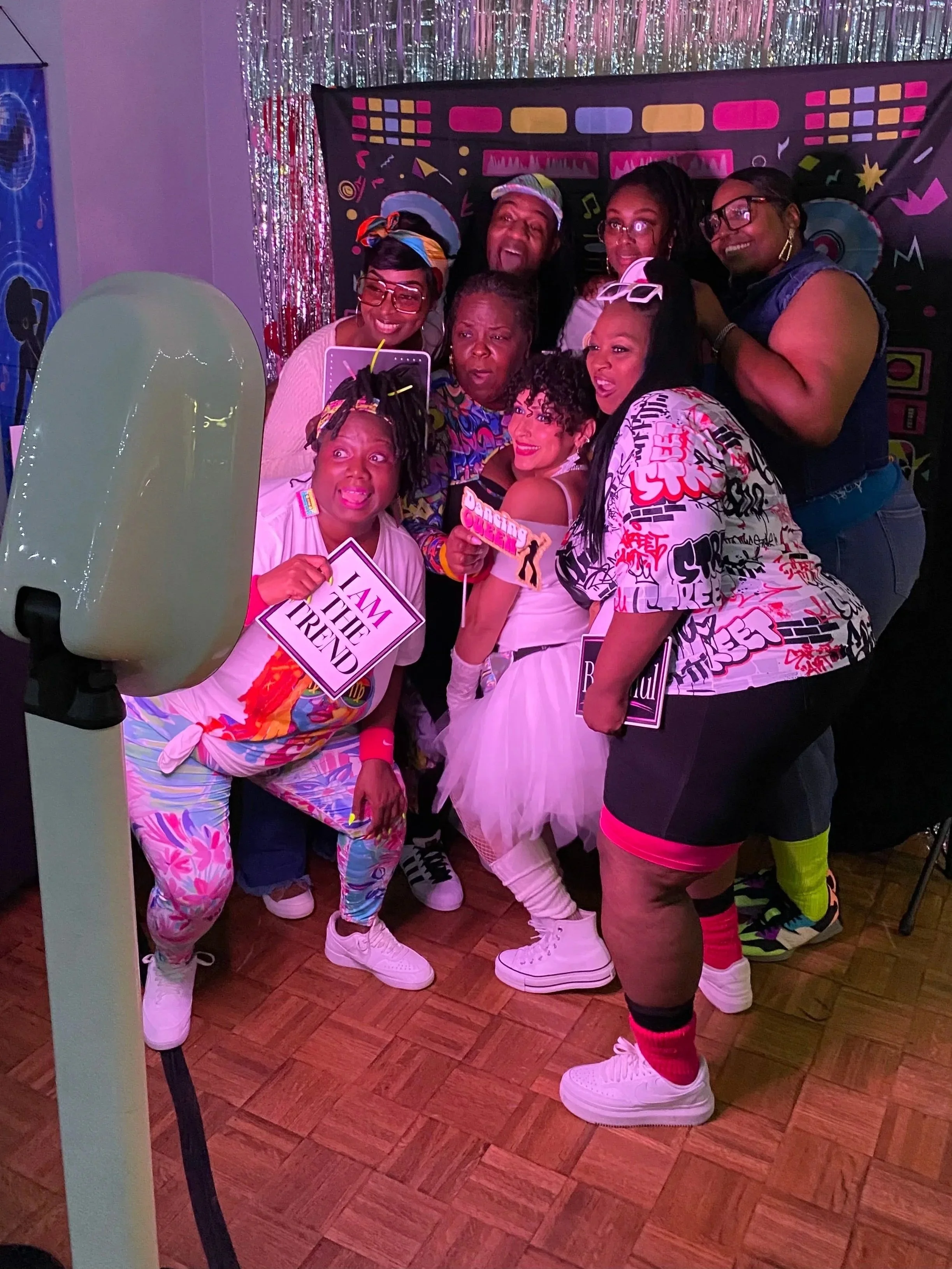 Group of six people dressed in colorful 80s-style outfits taking a selfie in a decorated party room with metallic curtains and a themed backdrop.
