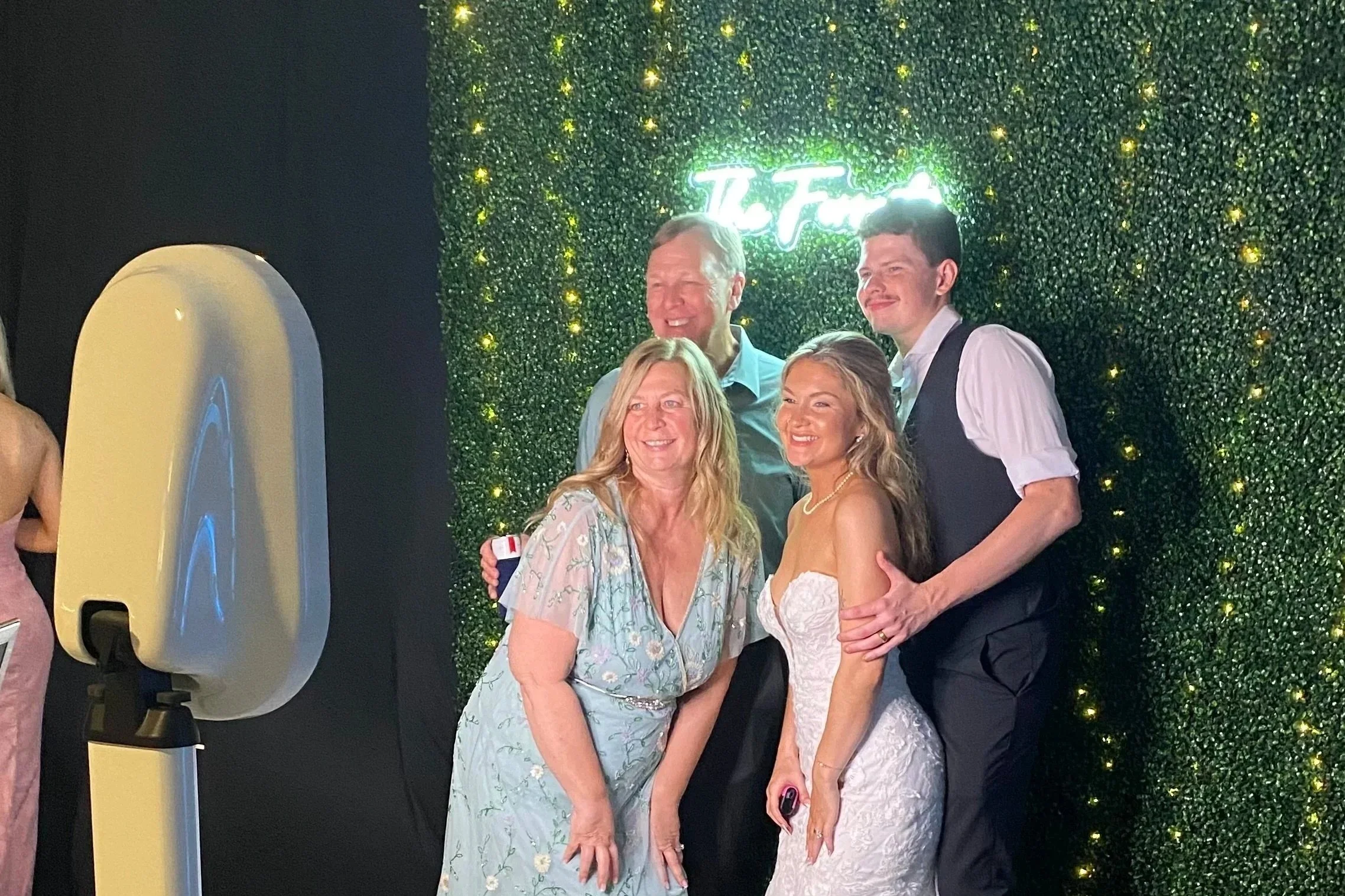 Group of five people dressed in formal attire, smiling and posing together in front of a green hedge wall with yellow fairy lights and a neon sign, at a celebration or event.