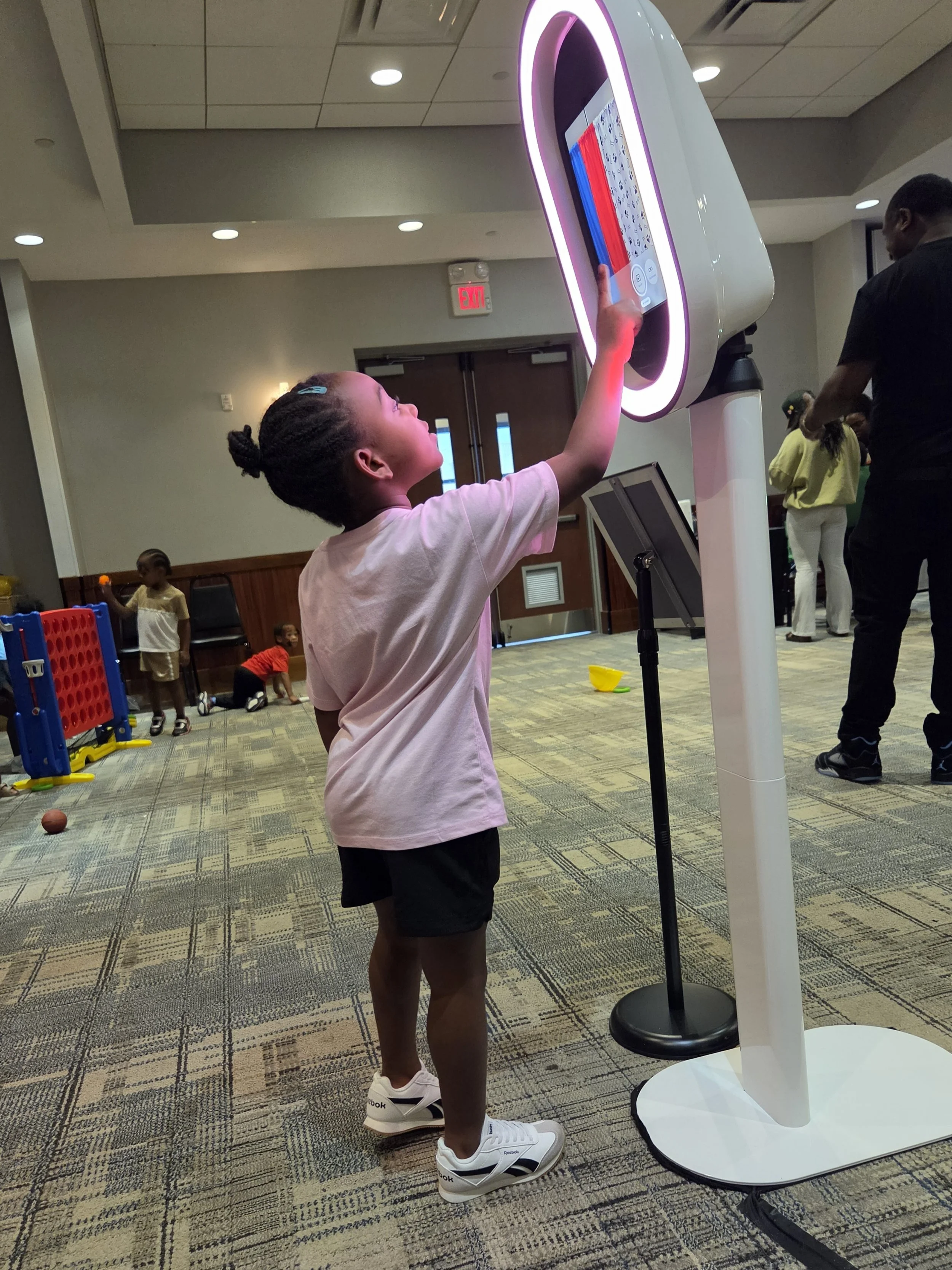 A young girl using a touchscreen kiosk at an indoor event, with other children and adults in the background.