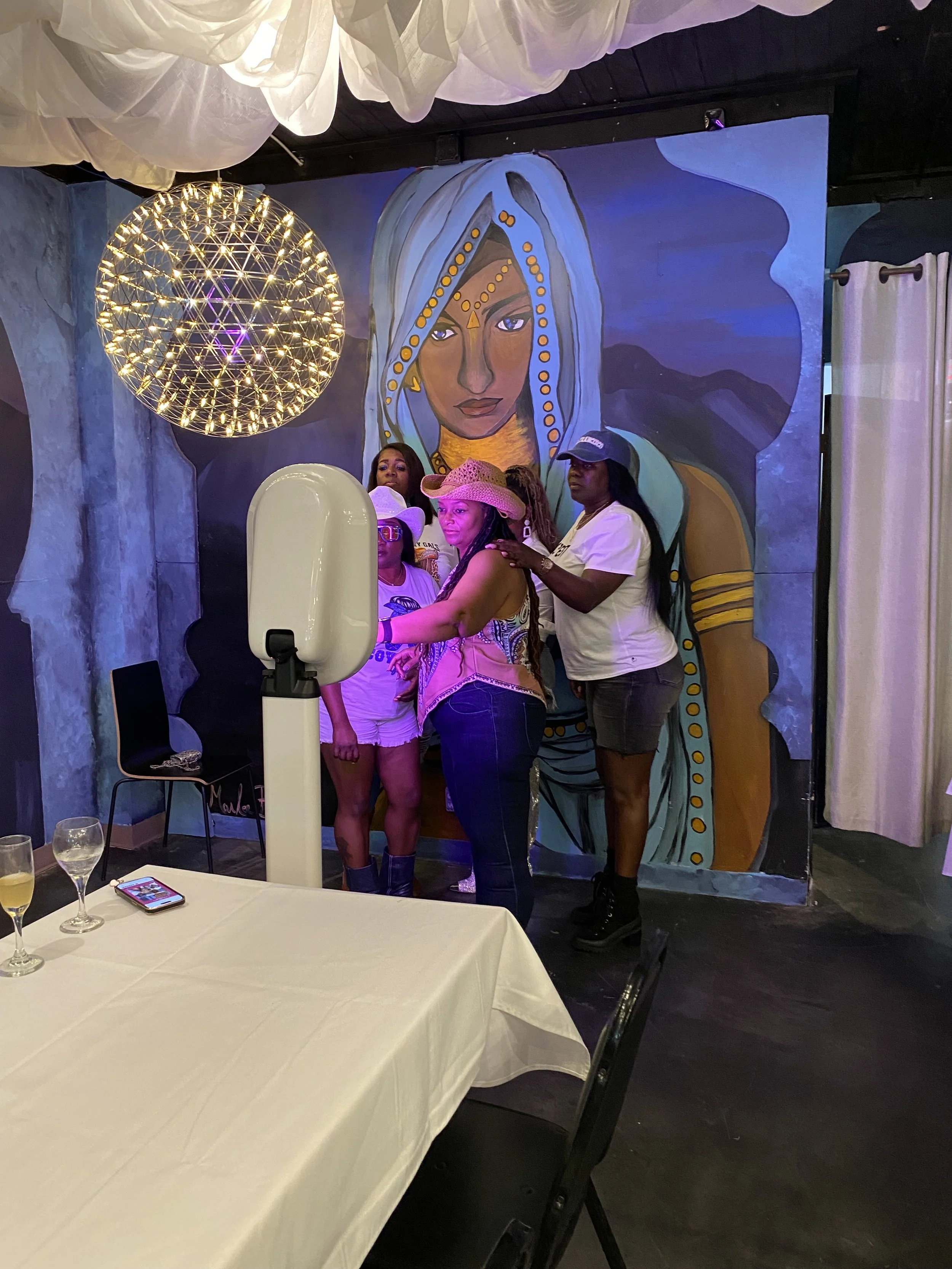 Four women standing in front of a mural of a woman's face with jewelry and a headdress, smiling while using PIXSBYIVORY PHOTO BOOTH.
