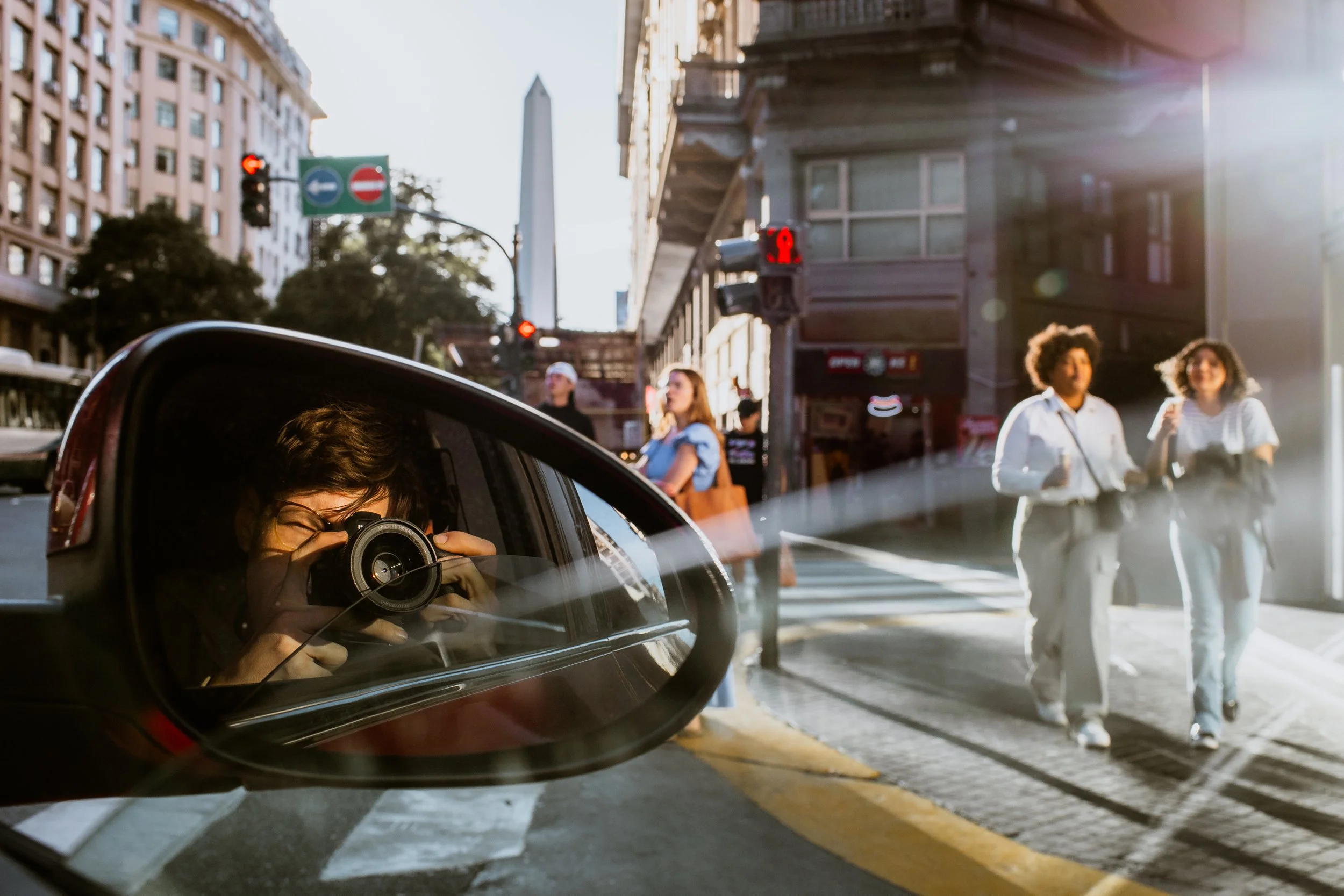Woman taking a photo with a camera, reflection visible in car side mirror, people walking on the street, traffic lights, and tall buildings in the city.