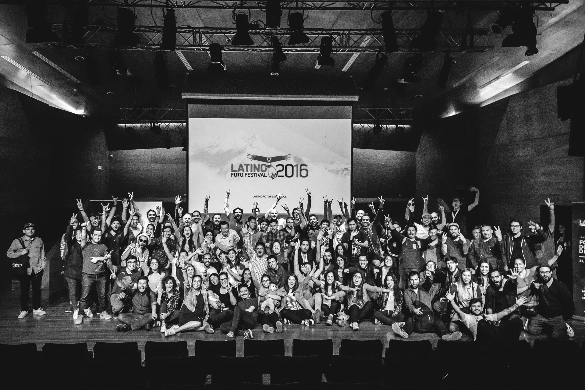 Group of people on stage at the Latino Foto Festival 2016, posing for a group photo in front of a large screen with the event logo, in a dimly lit auditorium.