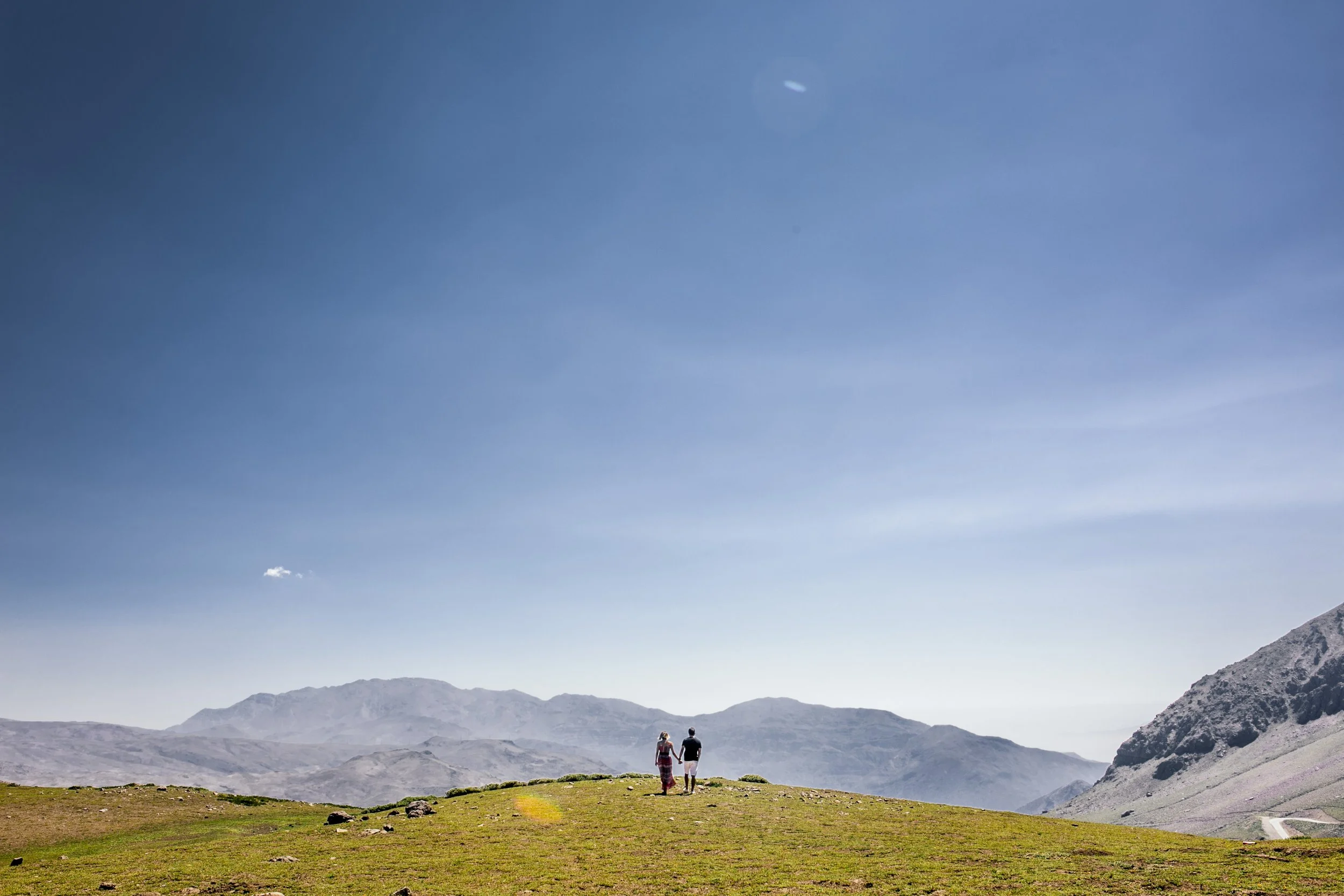 A couple walking hand-in-hand on a grassy hilltop with mountains in the background under a blue sky.