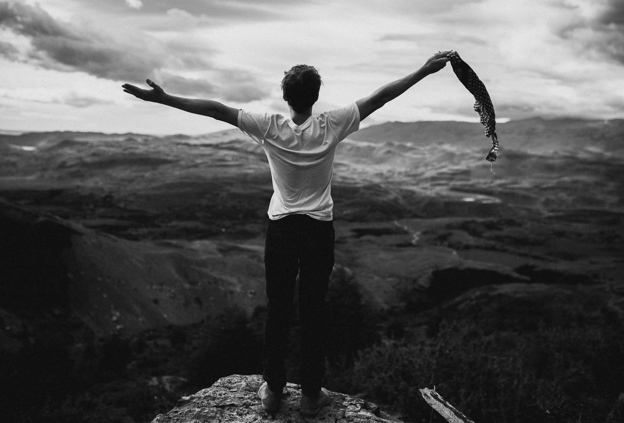 A person standing on a rock with arms outstretched, facing a vast landscape under cloudy skies, in black and white.