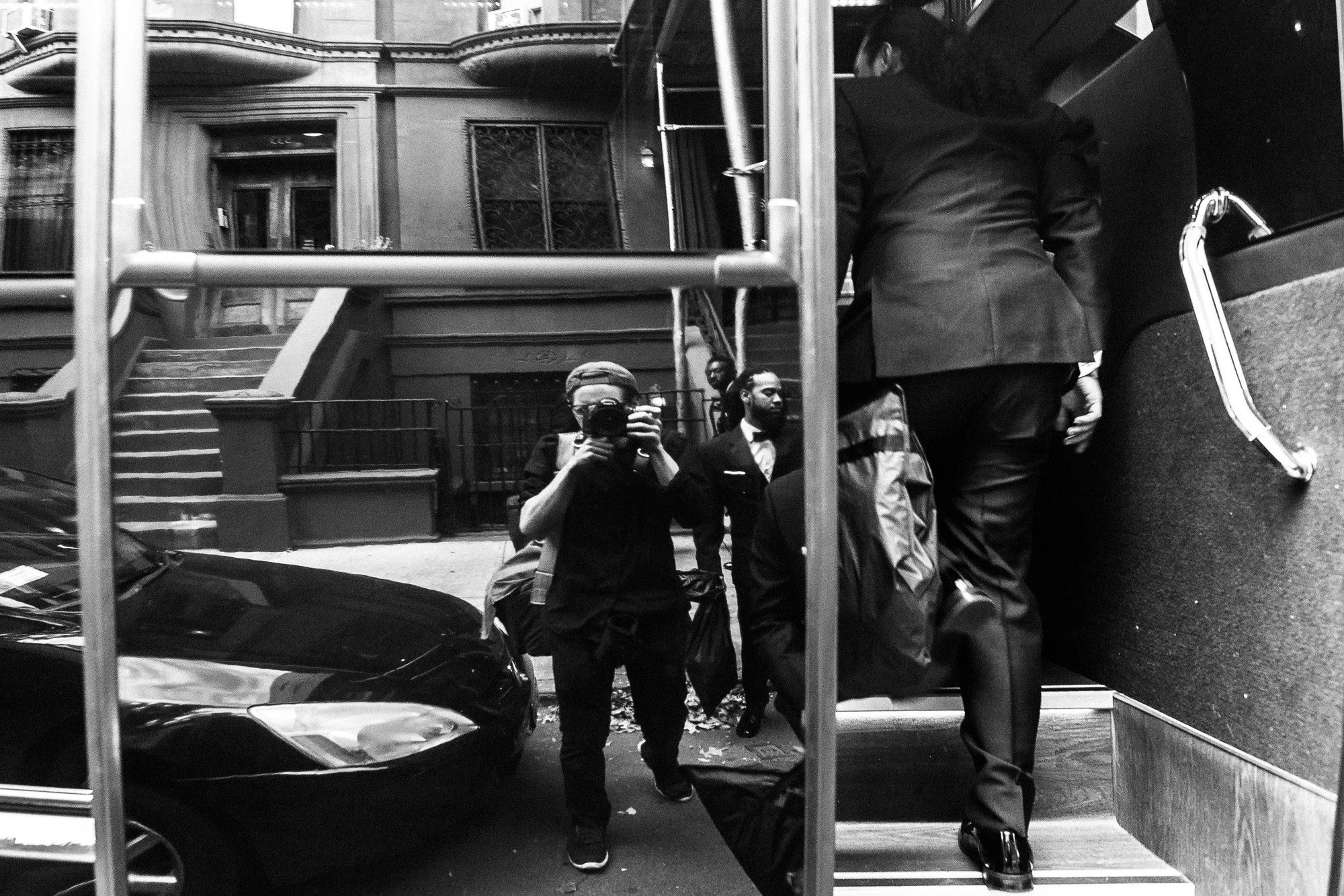 Black and white photo of a city scene, showing people walking on the sidewalk, cars parked along the street, and architecture of buildings in the background, captured through a window or mirror reflection.