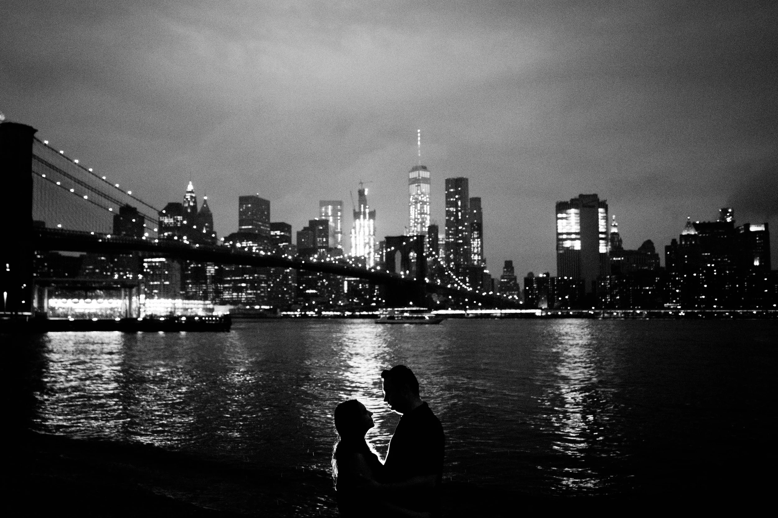 Silhouette of a couple embracing by the water with the Brooklyn Bridge and Manhattan skyline illuminated in the background at night, in black and white.