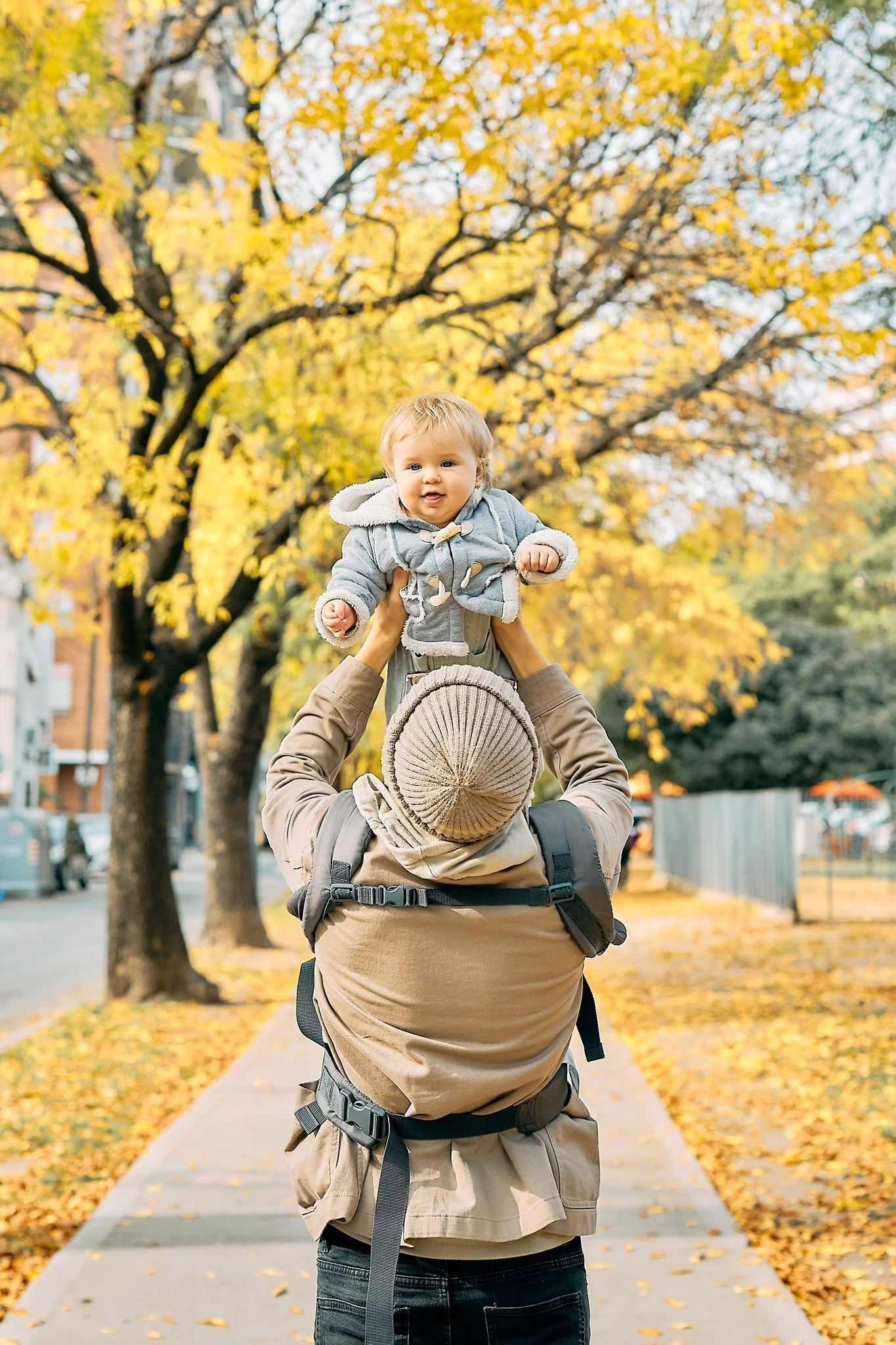 A child in a gray hoodie being held up by an adult with a green leaf backpack on, standing outdoors on a sidewalk with fallen yellow and orange leaves, and a large tree with yellow autumn leaves in the background.