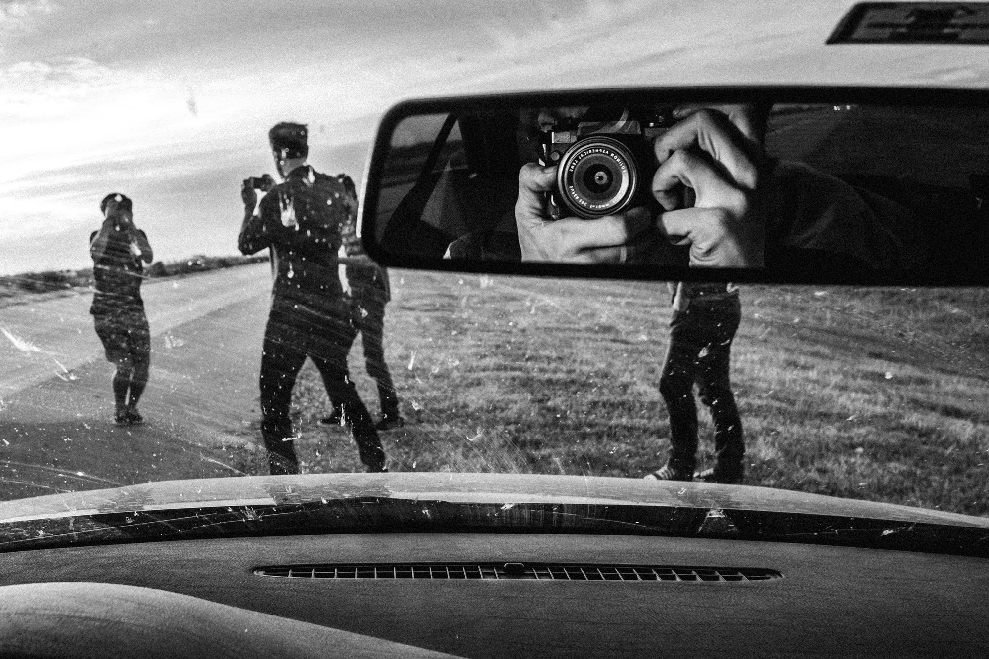 Black and white photograph taken from inside a car showing a rearview mirror with the reflection of a person holding a camera, taking a photo of two people standing on the roadside. One person is wearing a hat and jacket, and the other is wearing a j