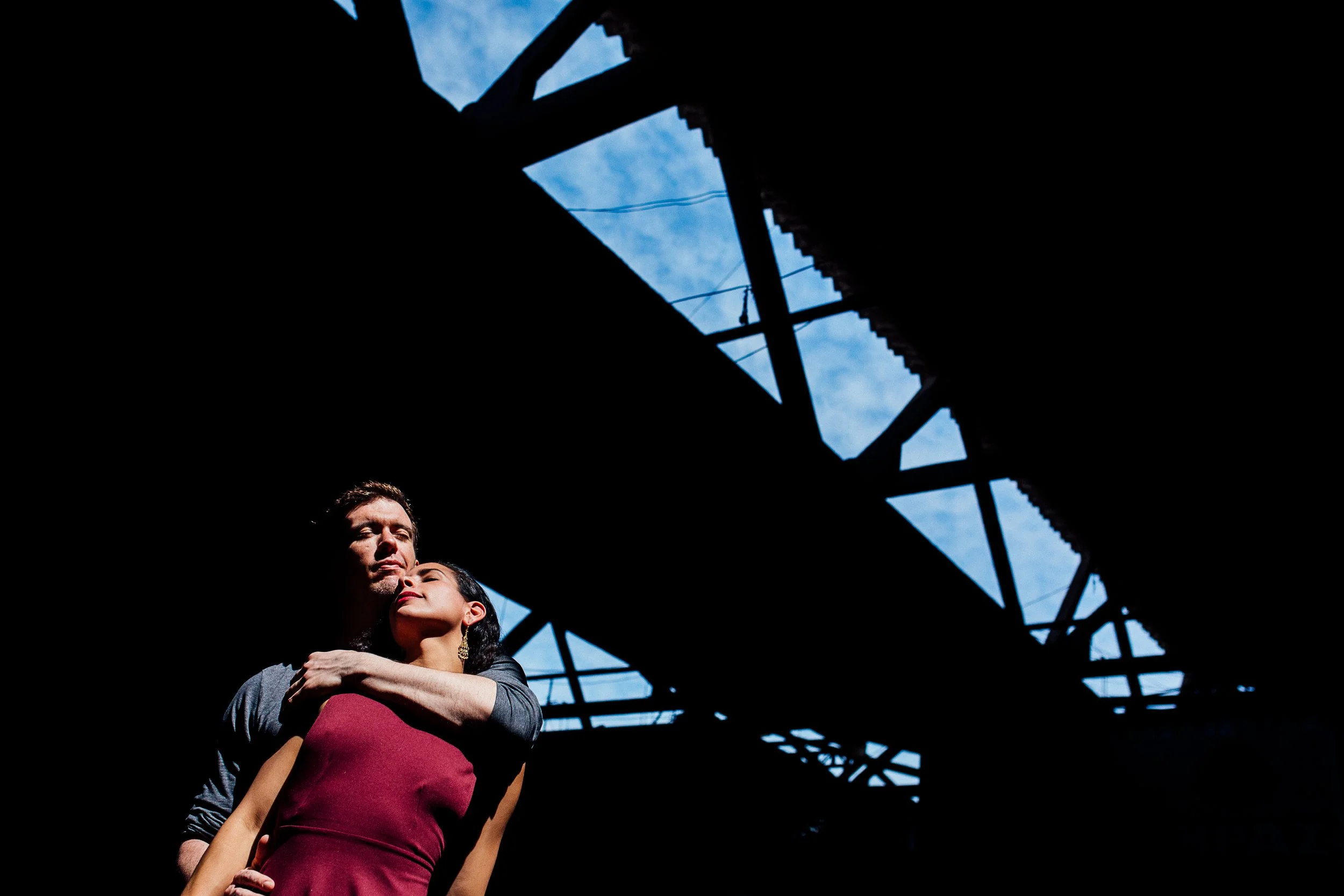 A man and woman embrace under an overpass with blue sky and clouds visible through the structural beams.