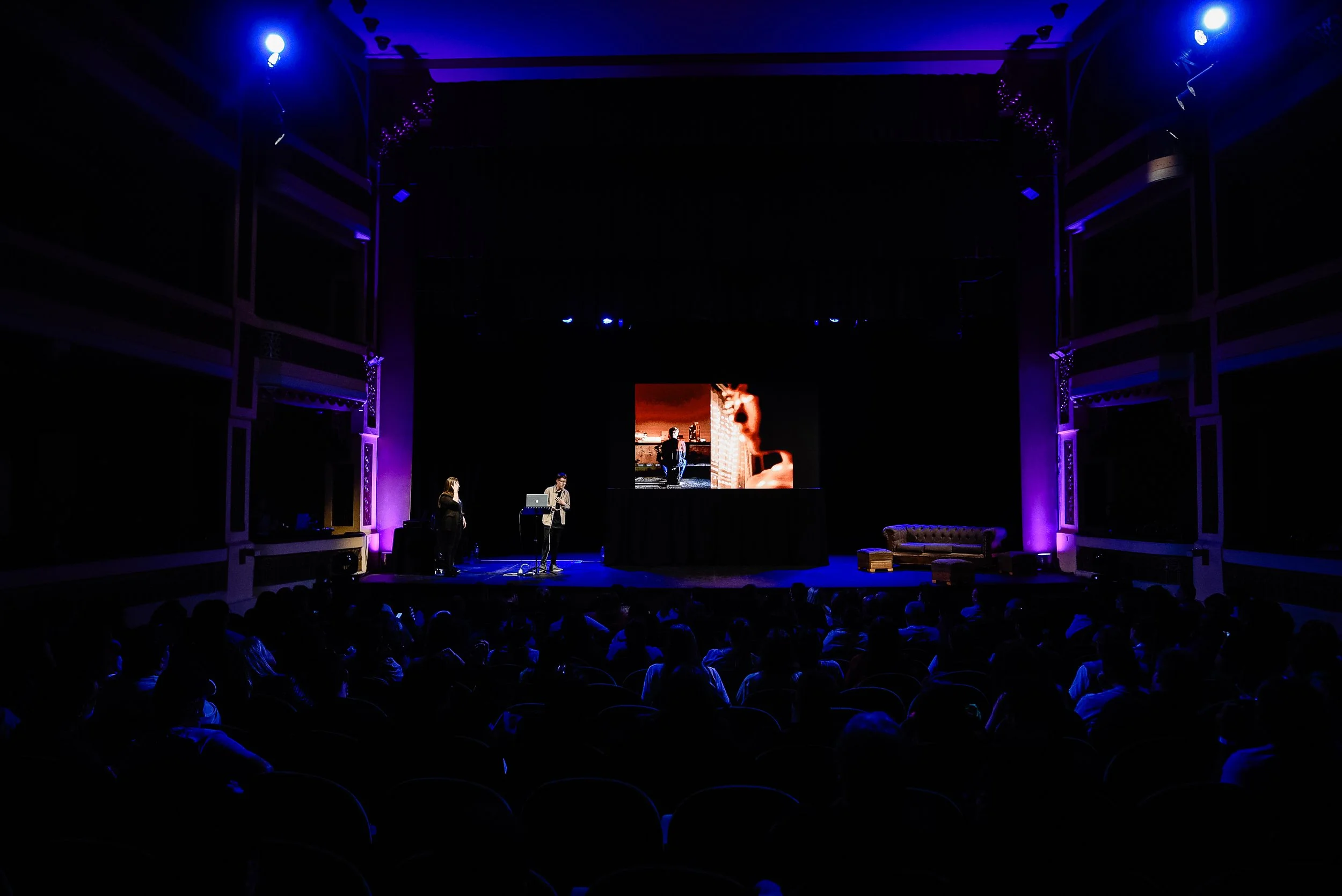 People on a stage in a dark auditorium with a large screen displaying a close-up of a person's face.