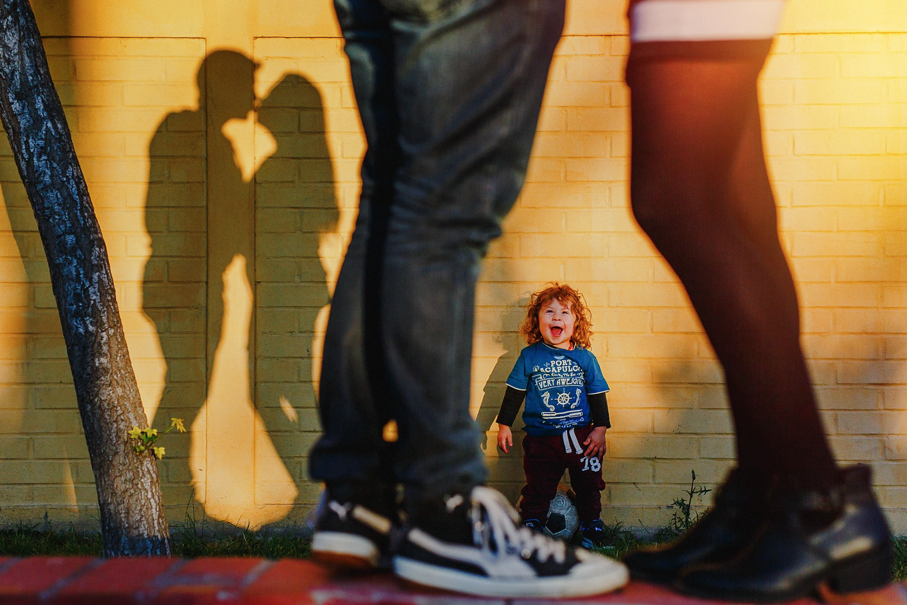 A young boy with red curly hair laughing and smiling, sitting against a yellow brick wall, with the shadows of two larger people in the foreground.