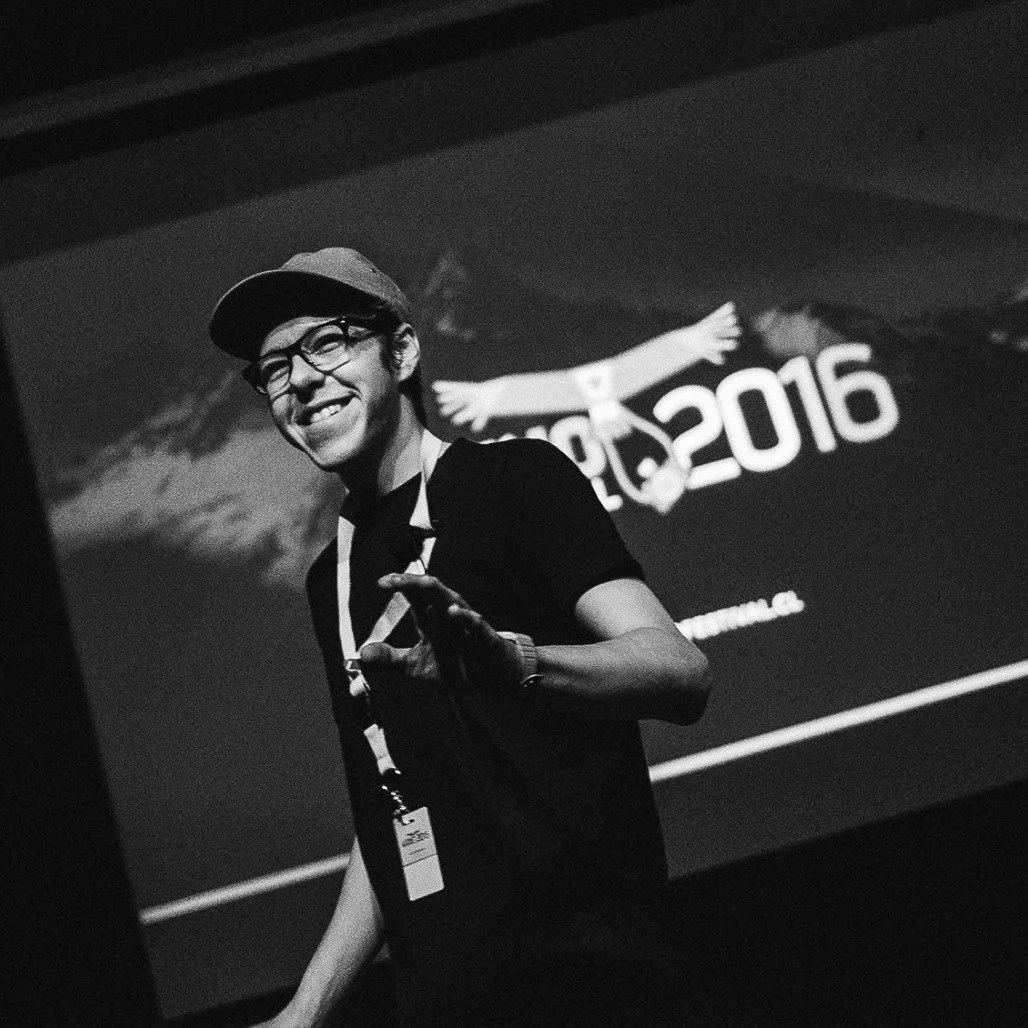 A young man wearing glasses, a hat, and a lanyard, smiling and making a peace sign, standing in front of a projection that reads '2016' and depicts a mountain and a person reaching out.