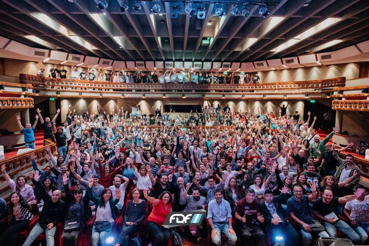 A large group of people in an auditorium with raised hands for a group photo, with some people on the upper balcony. The audience appears happy and enthusiastic.