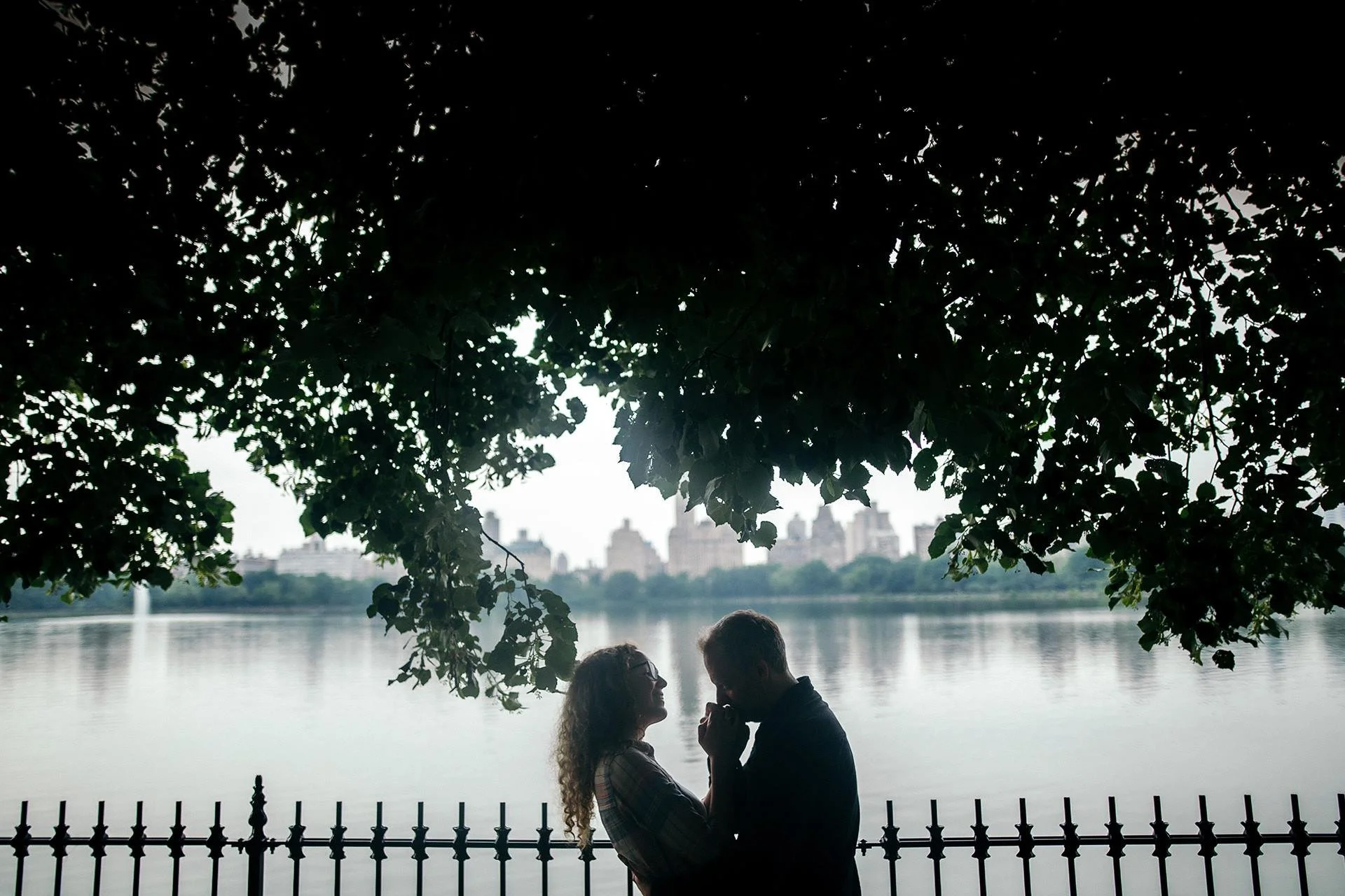 A couple stands close together by a fence near a lake, under a large tree with city buildings in the background, silhouetted against the bright sky.