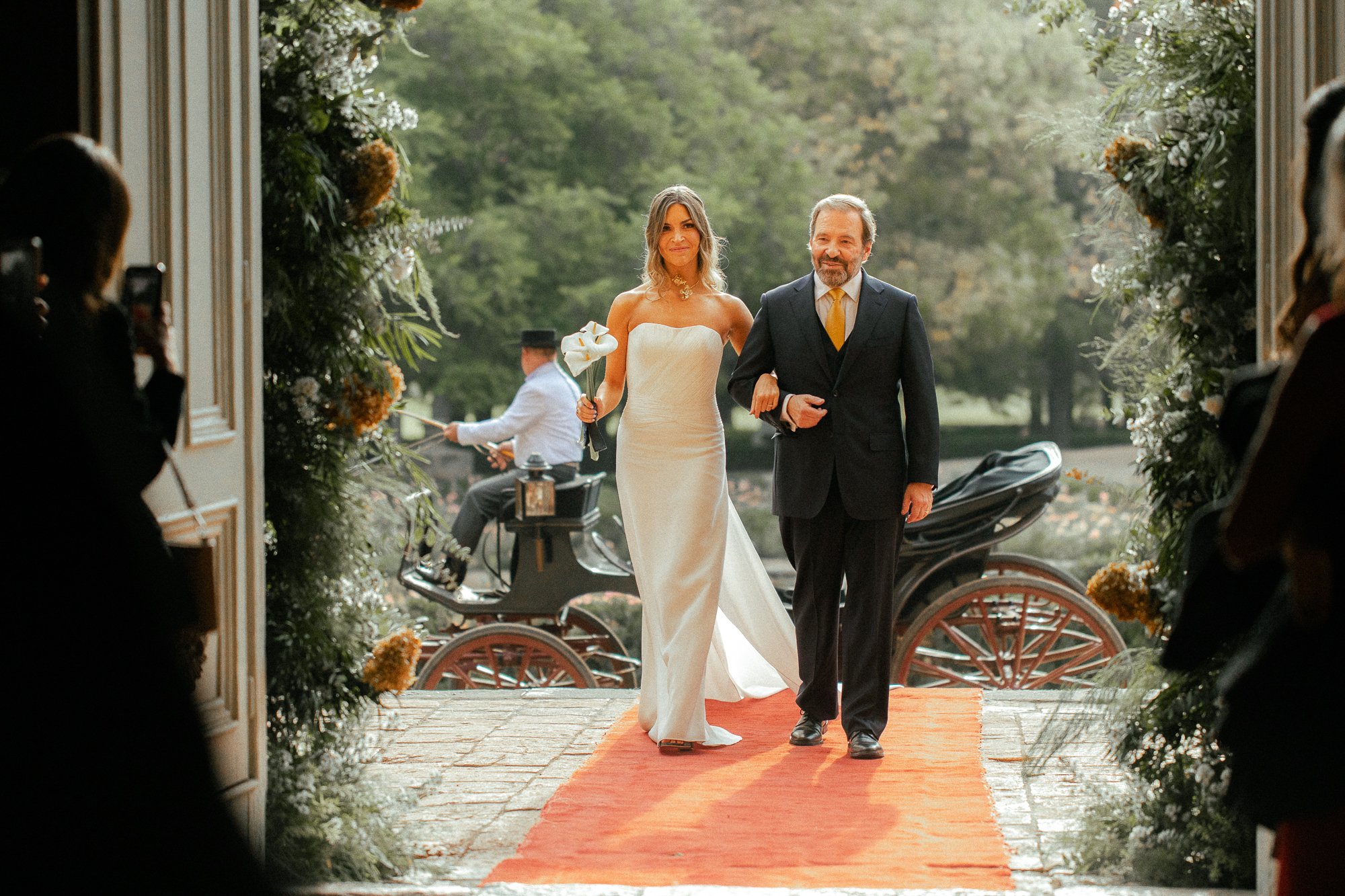 A bride in a white strapless wedding dress walking down the aisle with a man in a dark suit in an outdoor wedding ceremony. The aisle has a red carpet, and the background shows greenery and a horse-drawn carriage. Guests are taking photos on either s