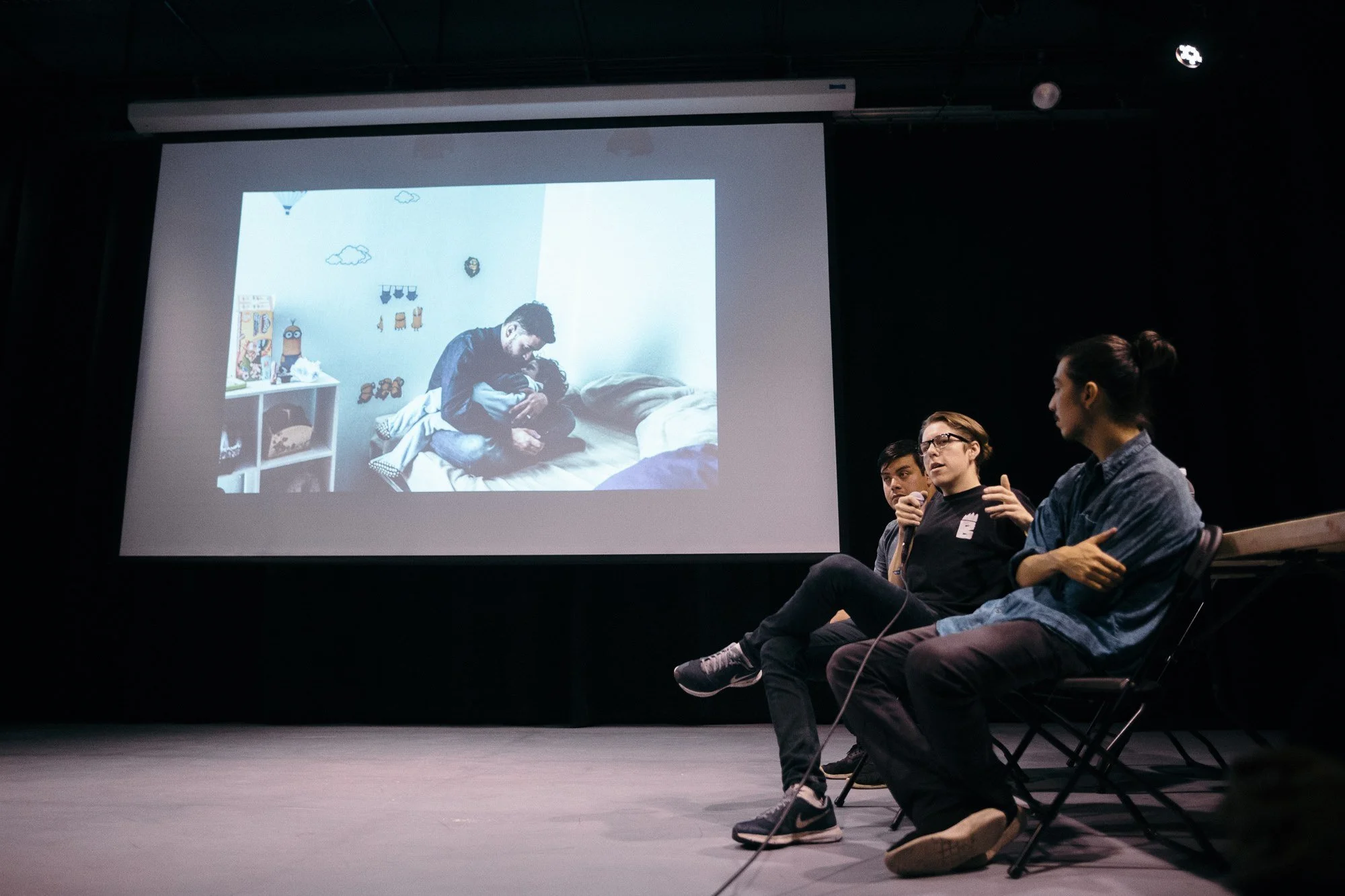 Three people are sitting on chairs in a dark auditorium, watching a presentation. A large screen displays a picture of a man hugging a child on a bed, with cartoon decorations on the wall.