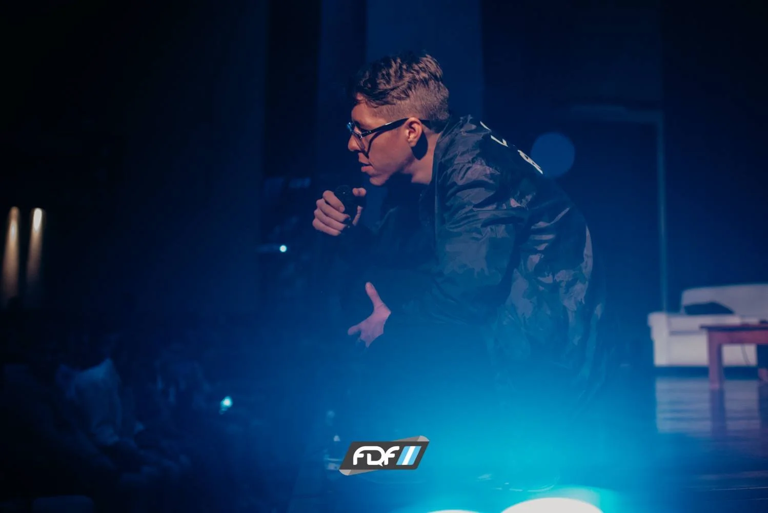 A man wearing glasses and a black jacket performing with a microphone on a dimly lit stage. There is a logo at the bottom with the letters 'FD' in white and blue.