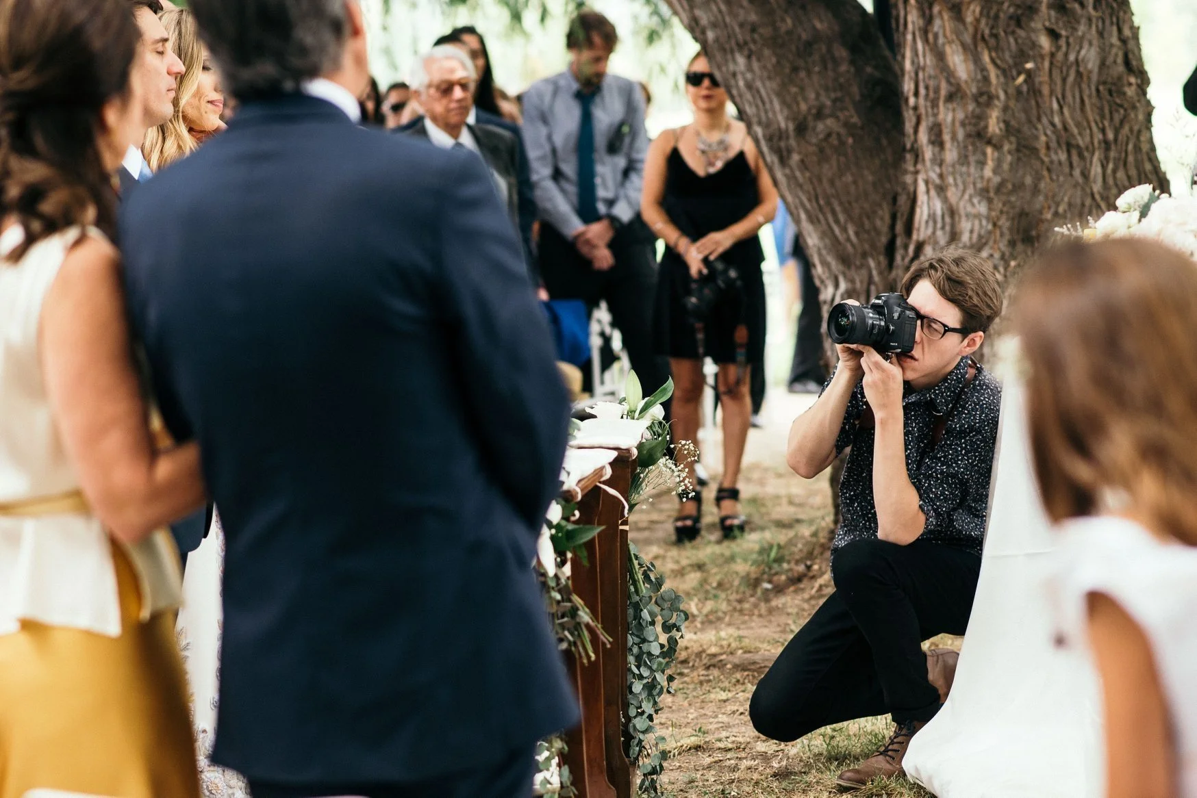 A photographer kneeling with a camera captures a wedding ceremony outdoors under a large tree, with guests standing behind the bride and groom.