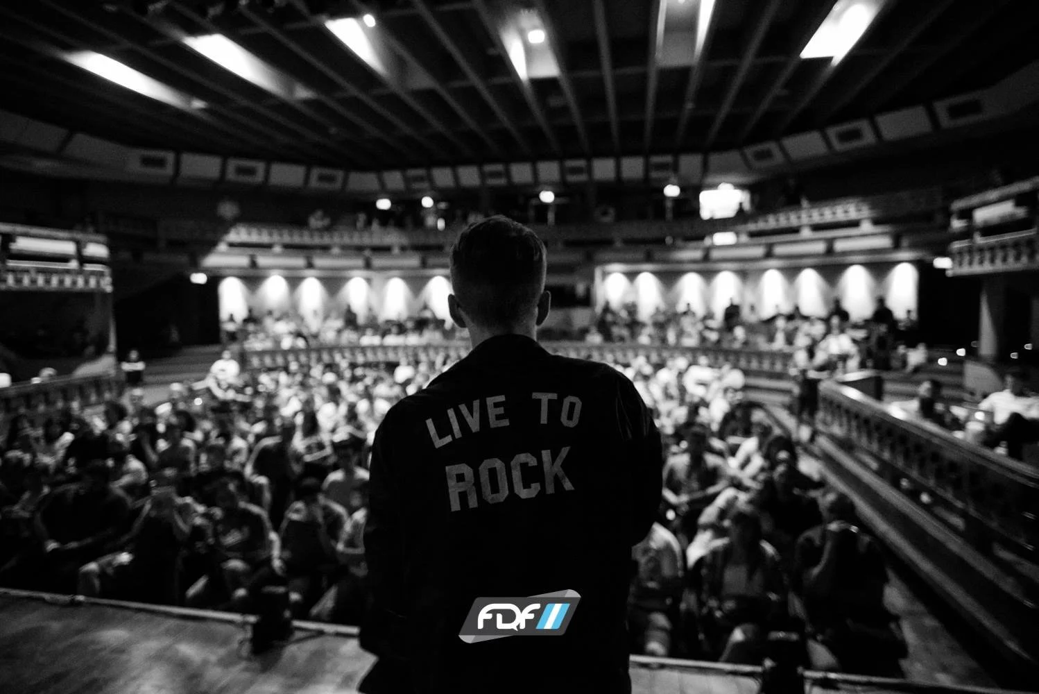 A person in a jacket with the text "Live to Rock" on the back is standing on a stage, facing a large audience in a theater or auditorium, with the audience seated and attentively watching a performance.