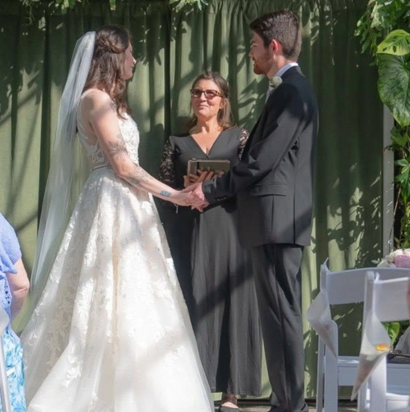 A bride and groom holding hands and looking at each other during their wedding ceremony, with an officiant standing between them and green foliage in the background.