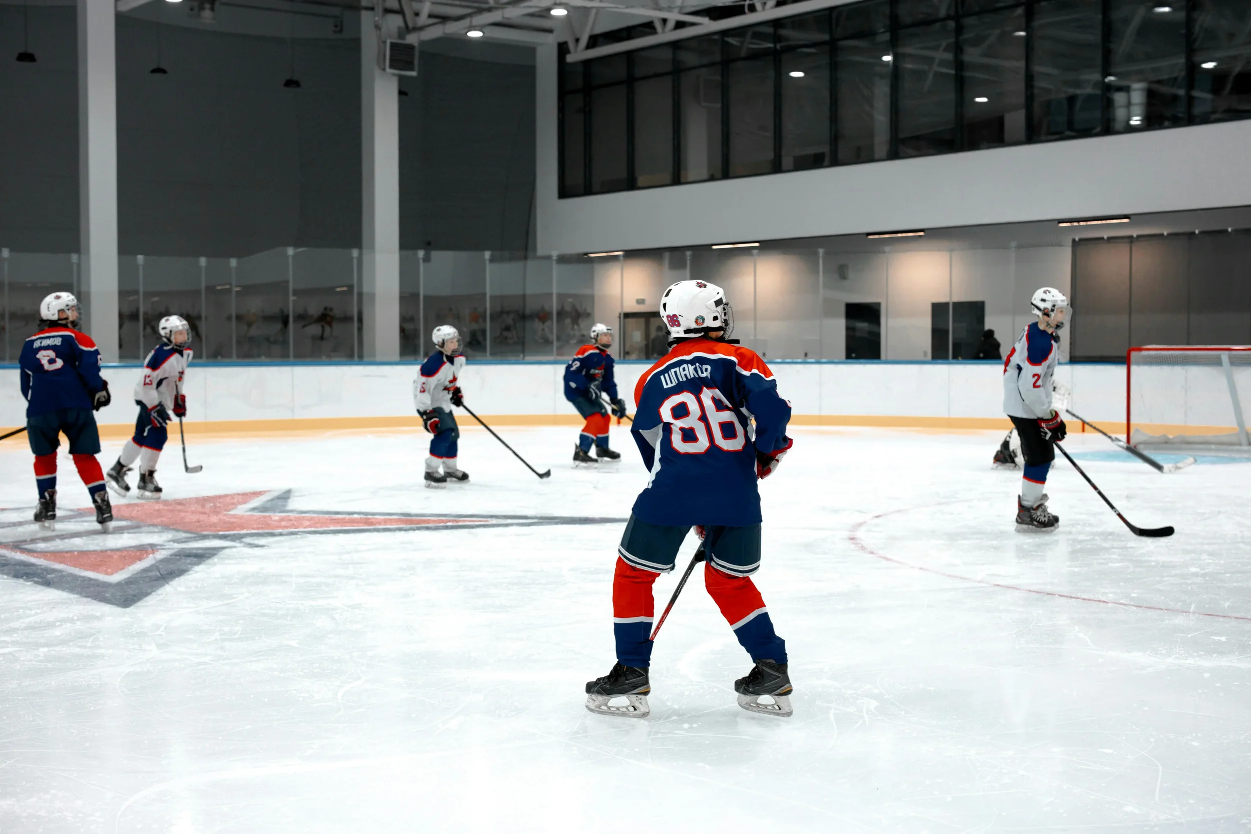 Hockey goalie in white, blue, and orange gear defending the goal, with a player in black approaching with a stick on ice rink.
