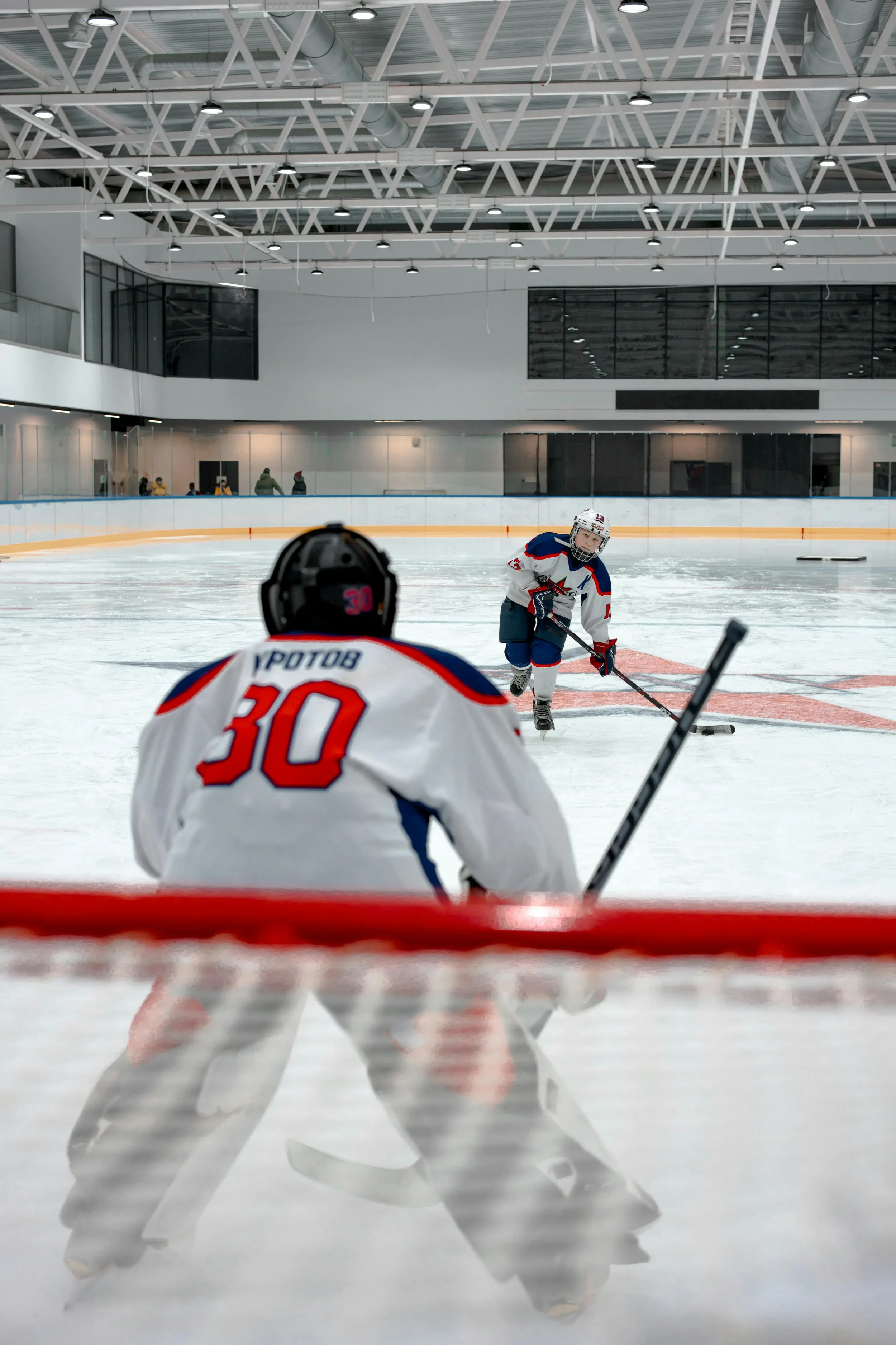 Youth hockey players in white and red jerseys playing on ice rink, with some players skating towards puck, while referees observe.