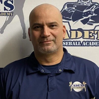 A man with a shaved head and goatee wearing a blue collared shirt with a Cadets Baseball Academy logo, standing in front of a grey background with the same logo.