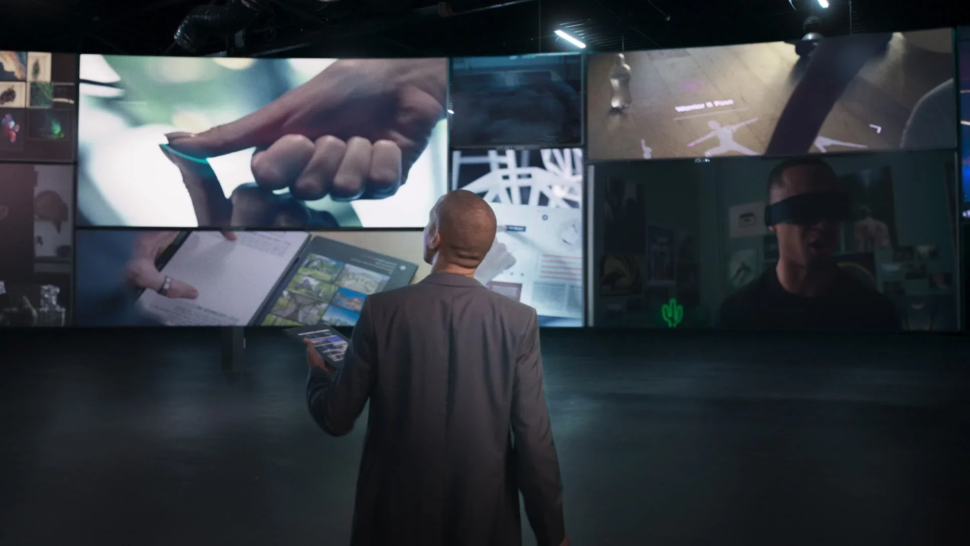 A man in a suit stands in front of multiple large screens displaying digital images, including a close-up of a hand touching a device, a person reading a book on a tablet, and a person wearing virtual reality goggles.