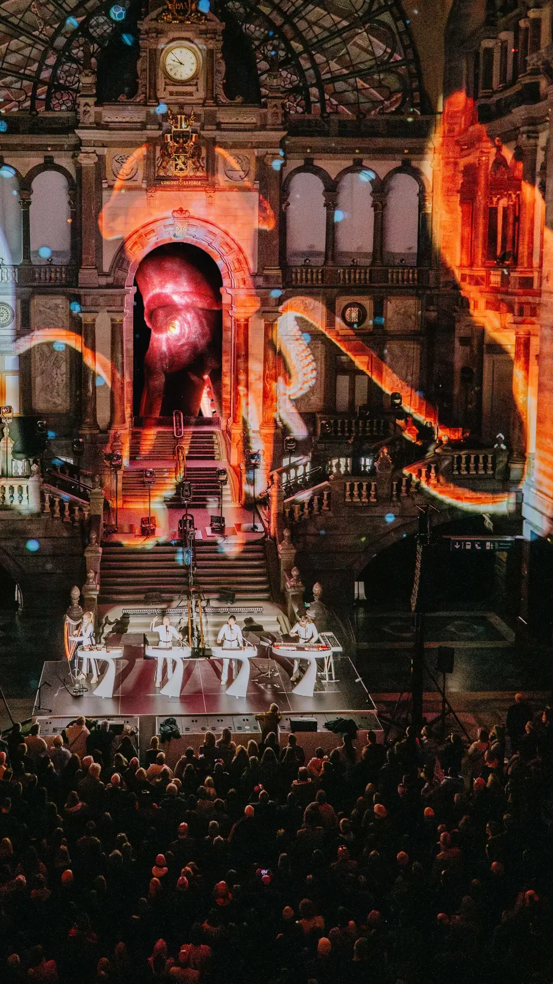 Live musical performance in a large ornate hall with projected visual effects on the backdrop