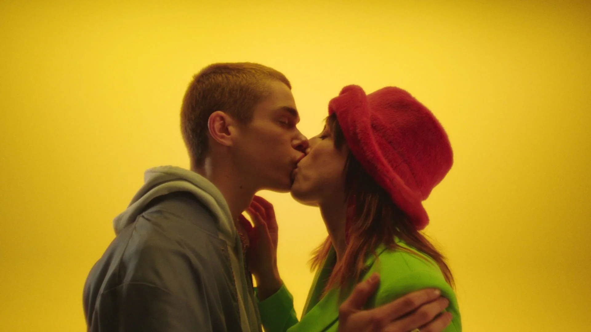 A young couple is kissing against a yellow background. The man has short hair and is wearing a gray hoodie, and the woman has long hair, is wearing a red hat and a bright green jacket.