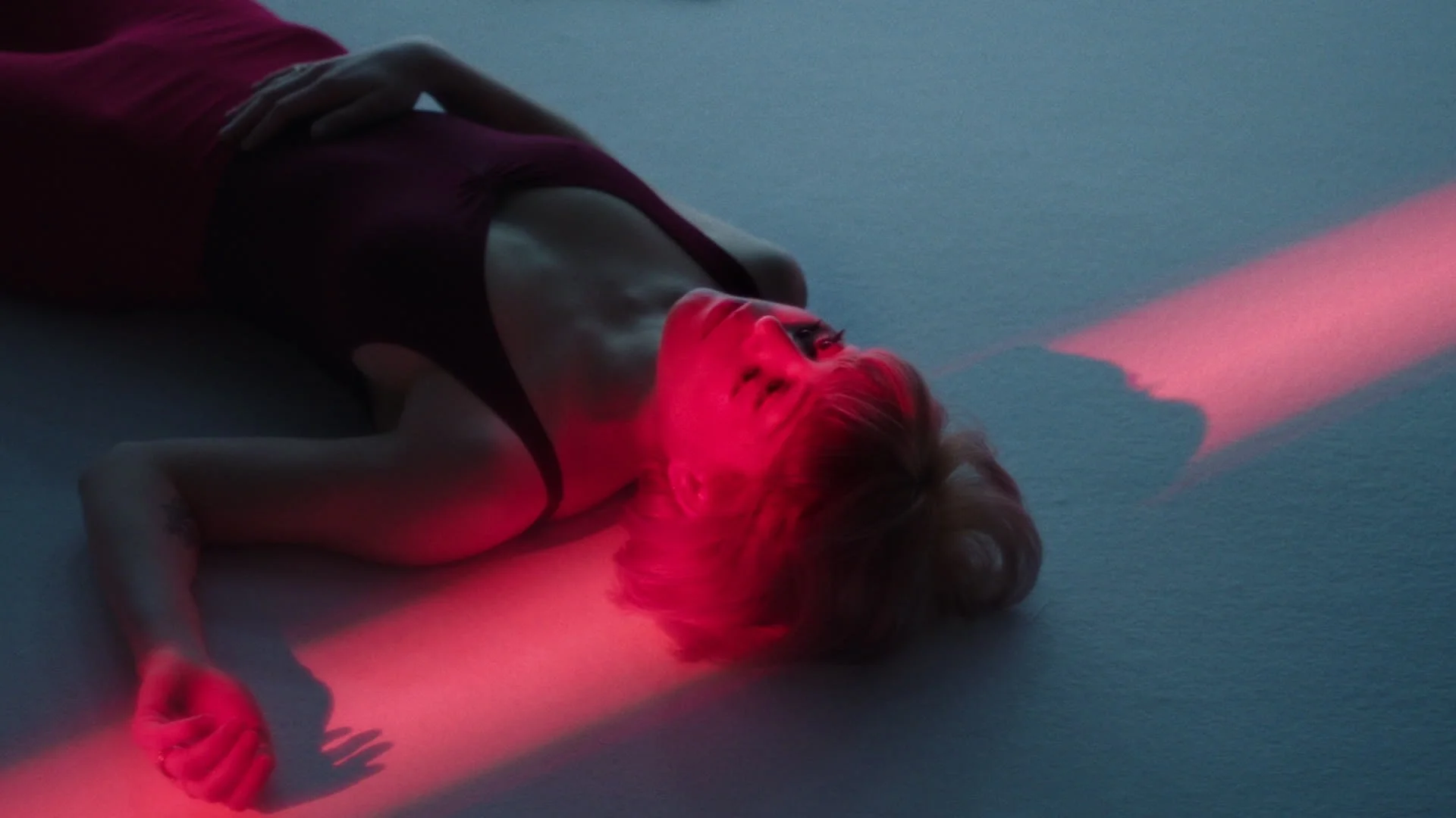 A woman with blonde hair lying on the floor, illuminated by red and blue lighting, wearing a sleeveless top, with one arm bent and one hand on her chest, looking up.