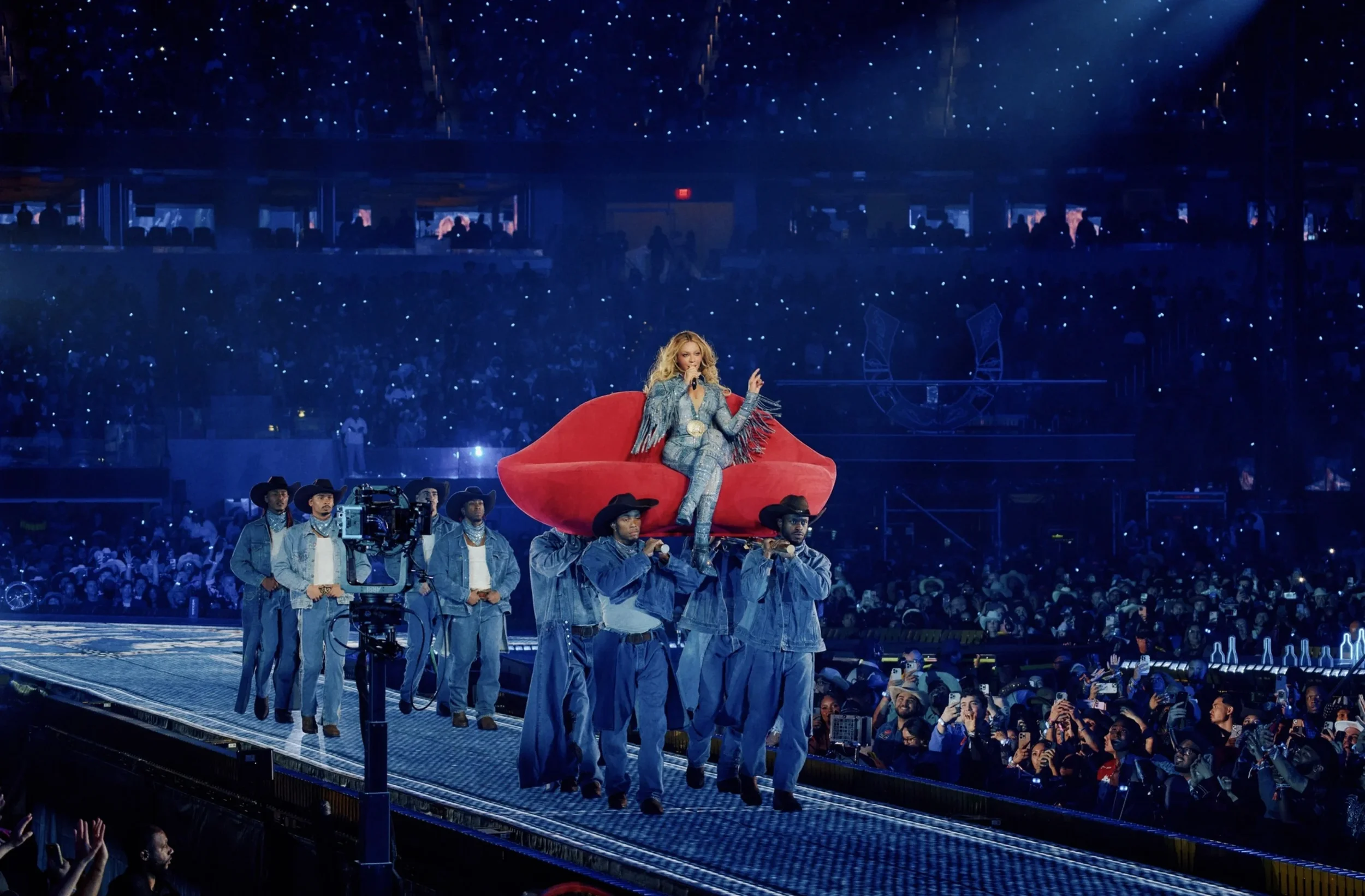 Country singer Taylor Swift performing on a giant red couch carried by crew members on a stage in front of a large audience at a concert.