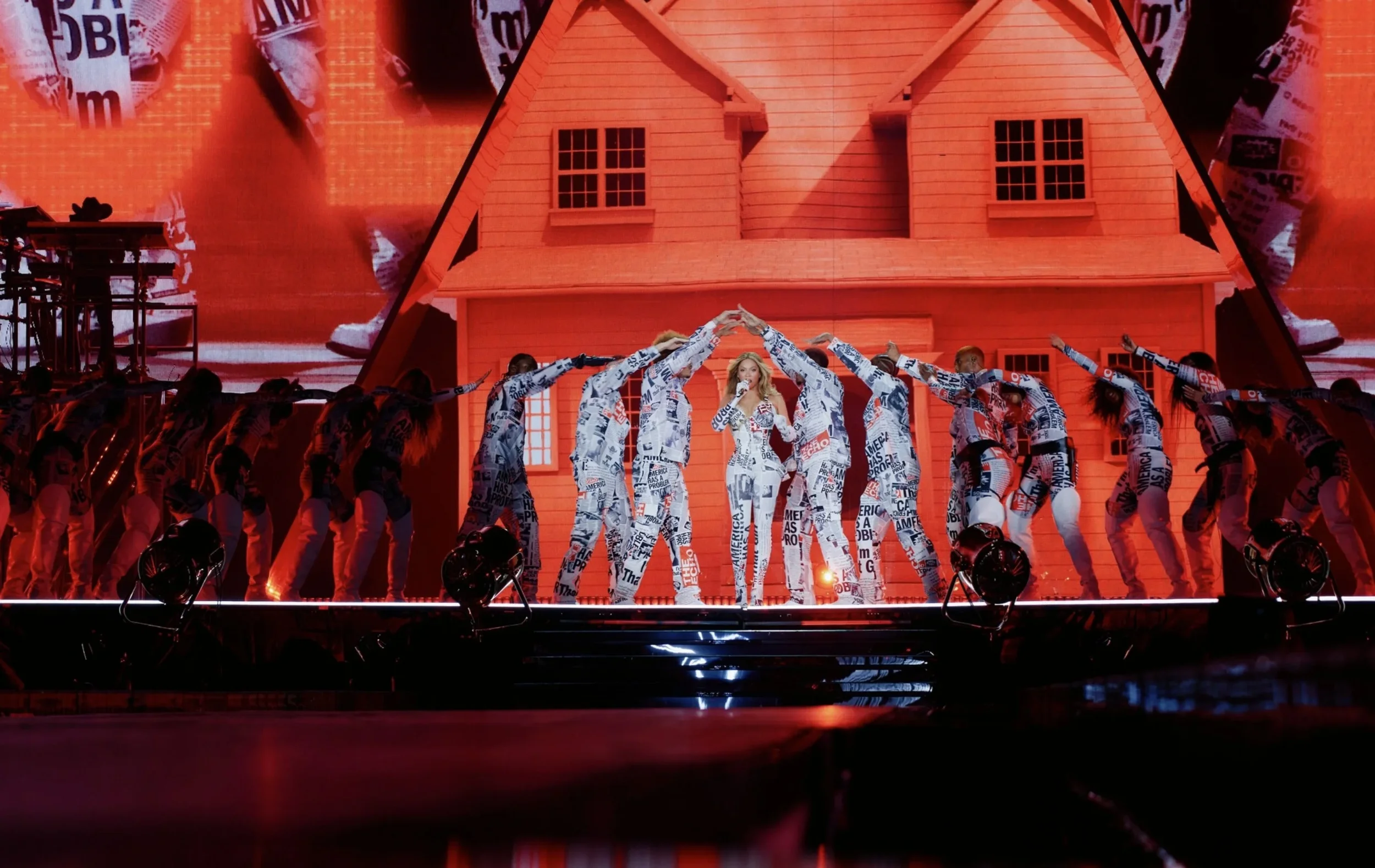 A performer on stage dressed in a newspaper print outfit singing into a microphone, surrounded by dancers also in newspaper print costumes, in front of a large red house backdrop.
