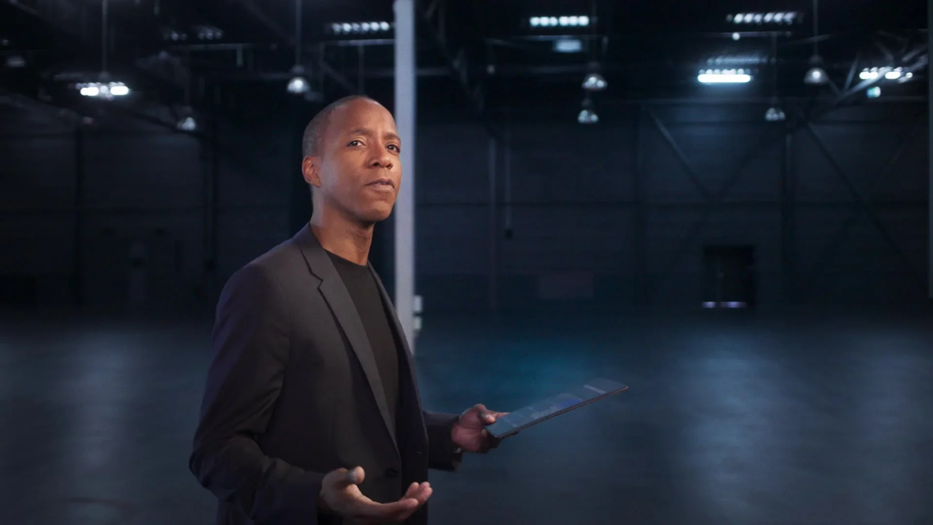 A man in a black suit holding a tablet in a large, empty, dark indoor space with high ceilings and lighting fixtures.