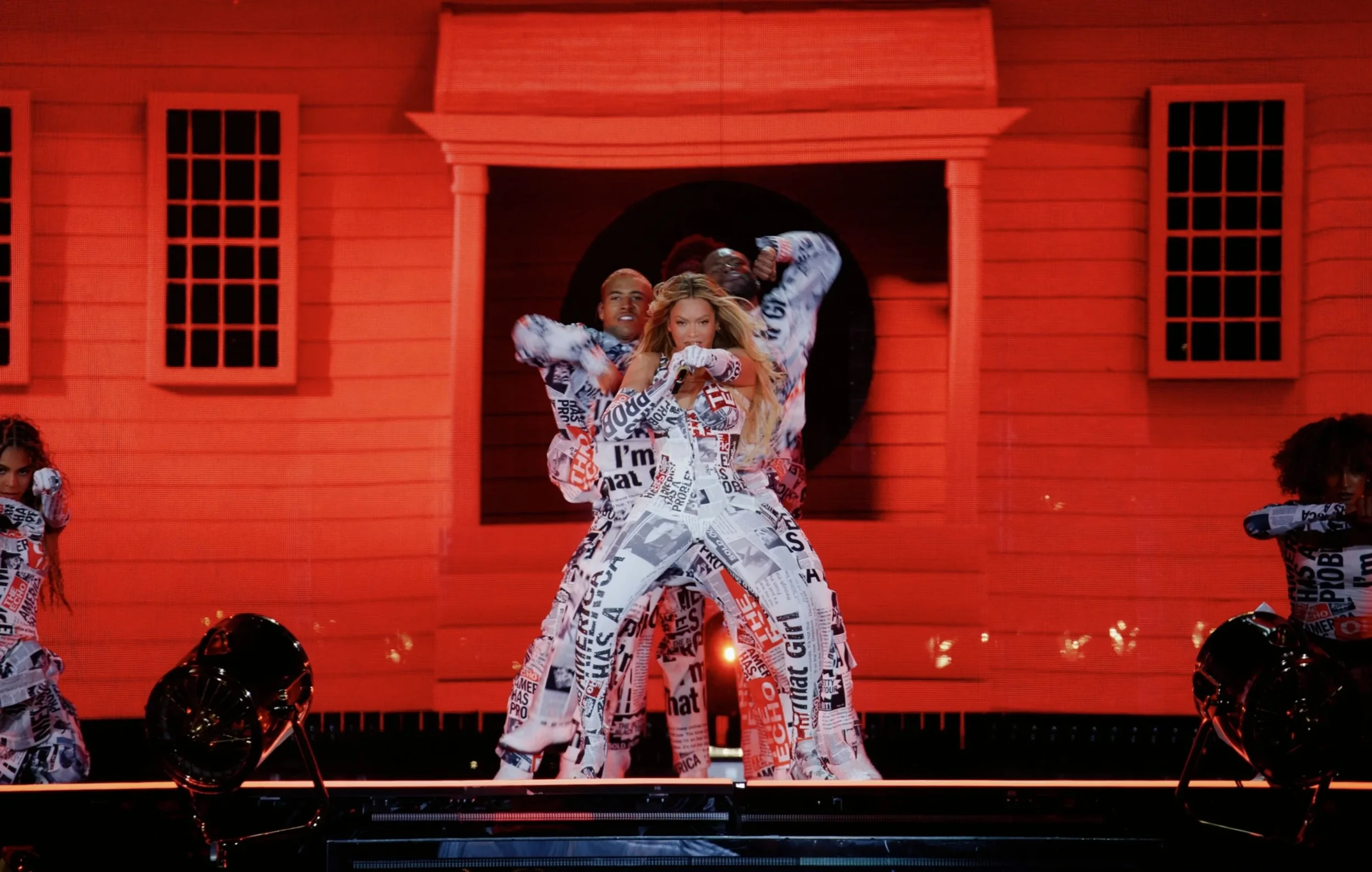 Pop singer perform on stage with backup dancers, all wearing newspapers print outfits, with a large red house prop in the background.