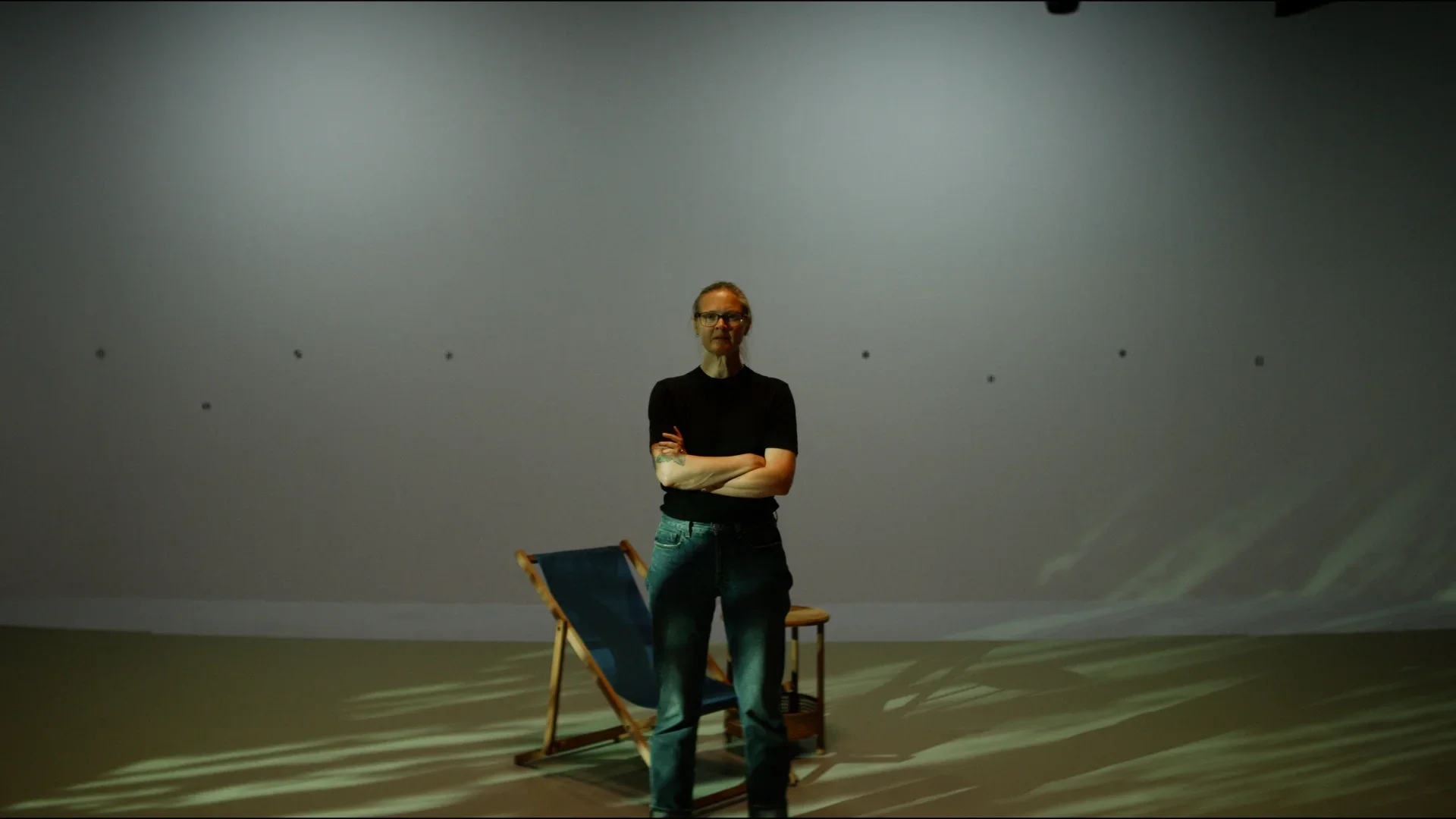 A woman standing with crossed arms in an empty room with a blank wall, a folding chair, and a small stool, illuminated by overhead lighting.