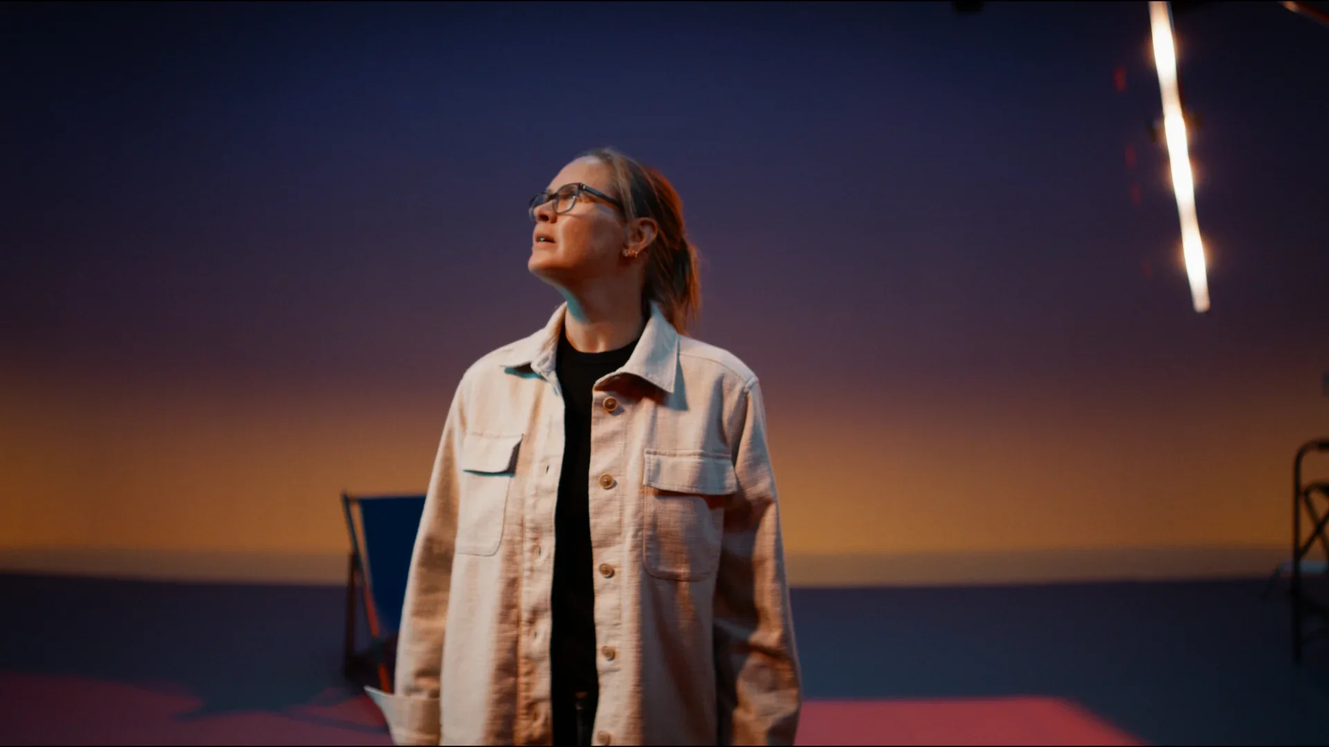 A woman with glasses and a denim jacket looking up at the sky with fireworks or streaks of light in the dark background during sunset or night.