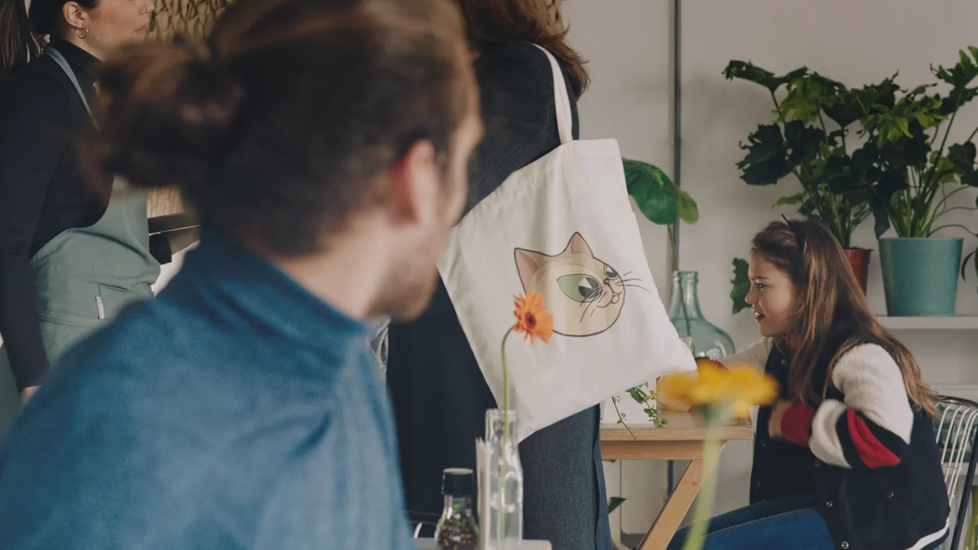 A young girl engaging with someone at an indoor gathering, with plants and decorative items on the background, and a tote bag with a cat illustration hanging on a person in the foreground.